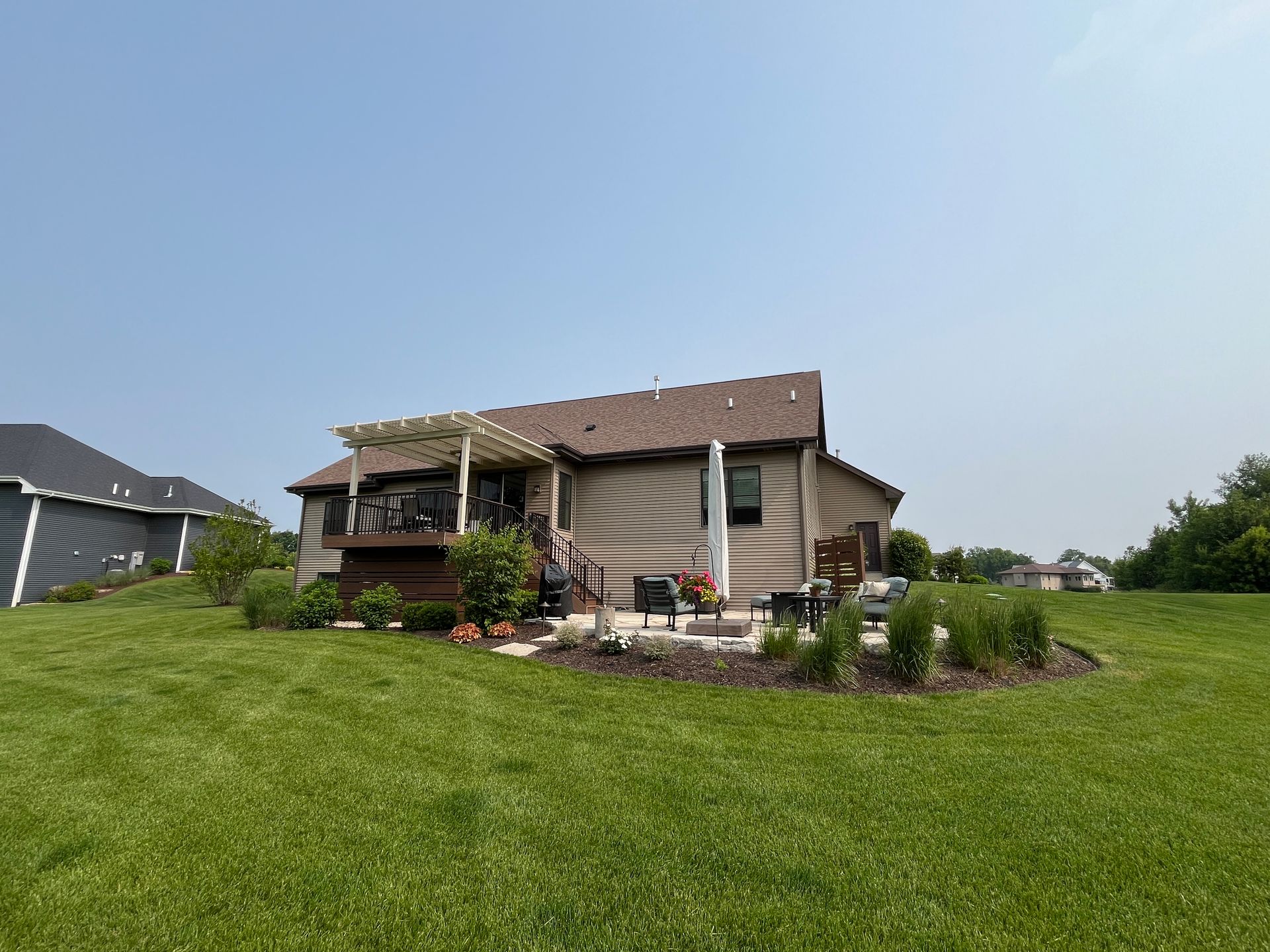 Backyard with house; patio furniture, deck with pergola, and green lawn under a blue sky.