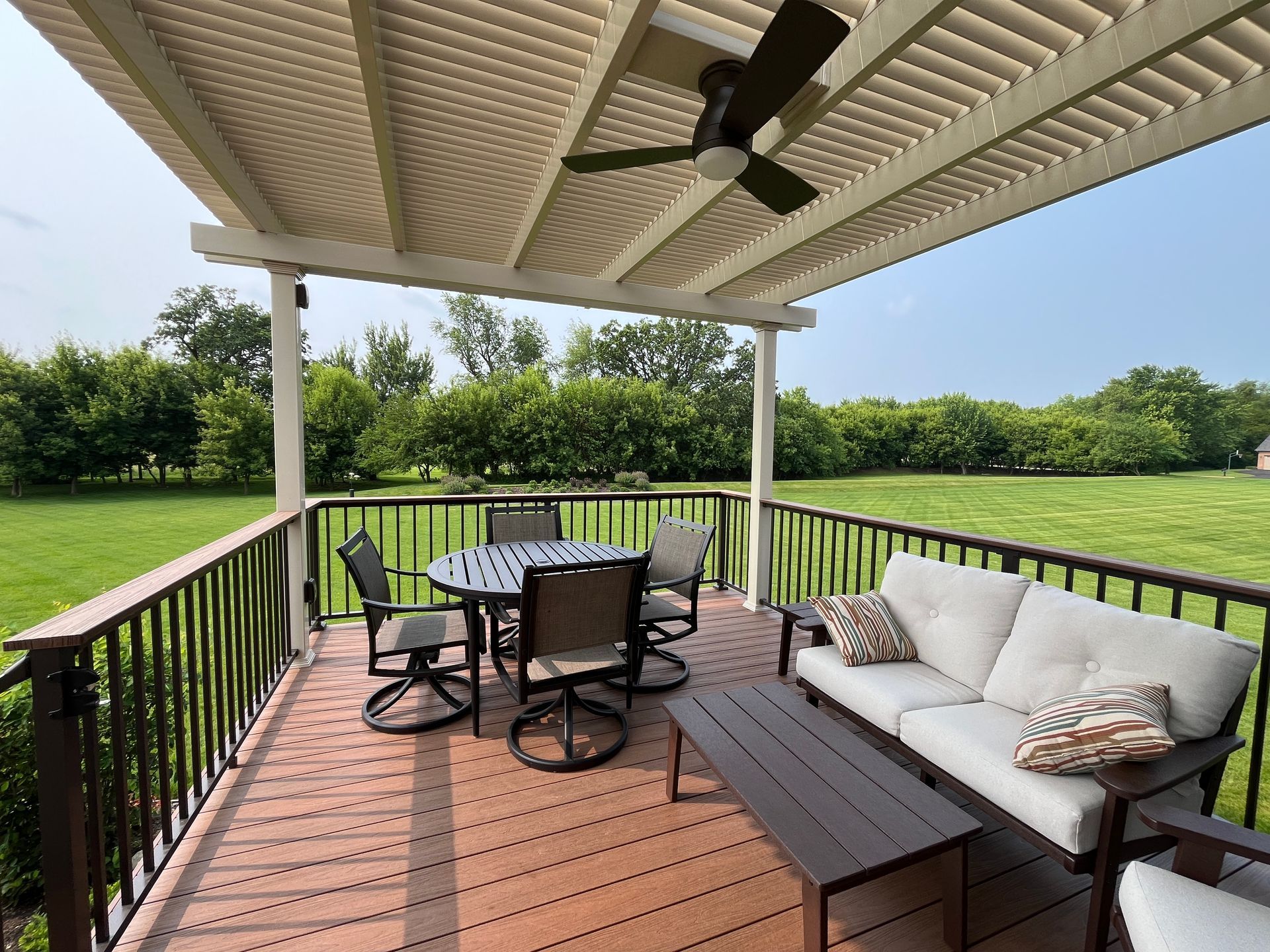 Covered deck with outdoor furniture: table and chairs, sofa, dark wood railing, and ceiling fan. Green lawn in the background.