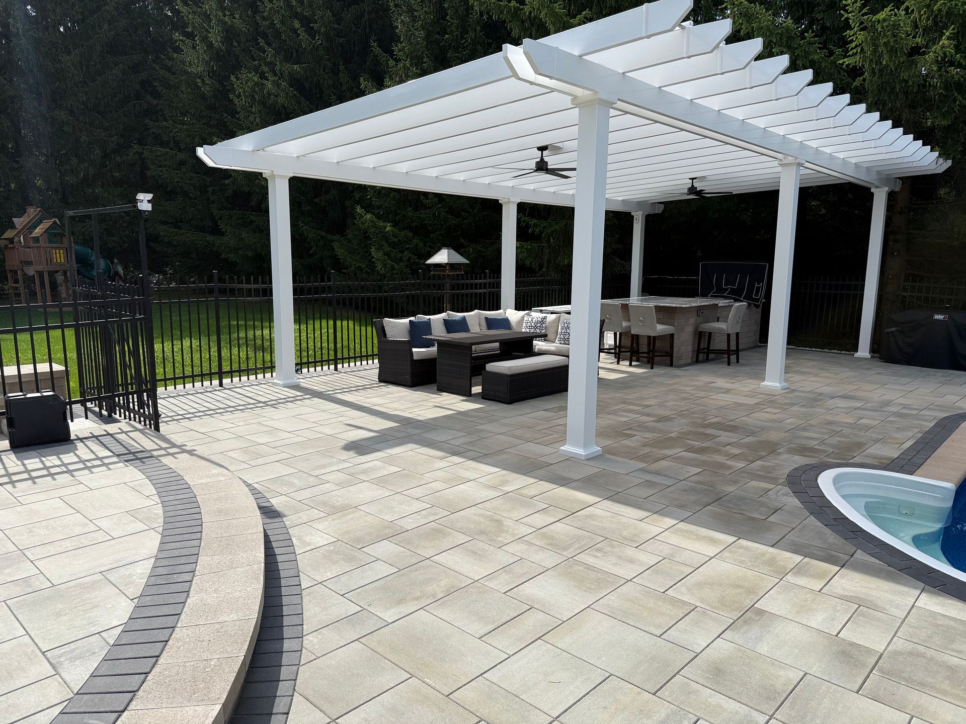 White pergola over patio with outdoor seating near a pool and black fence.