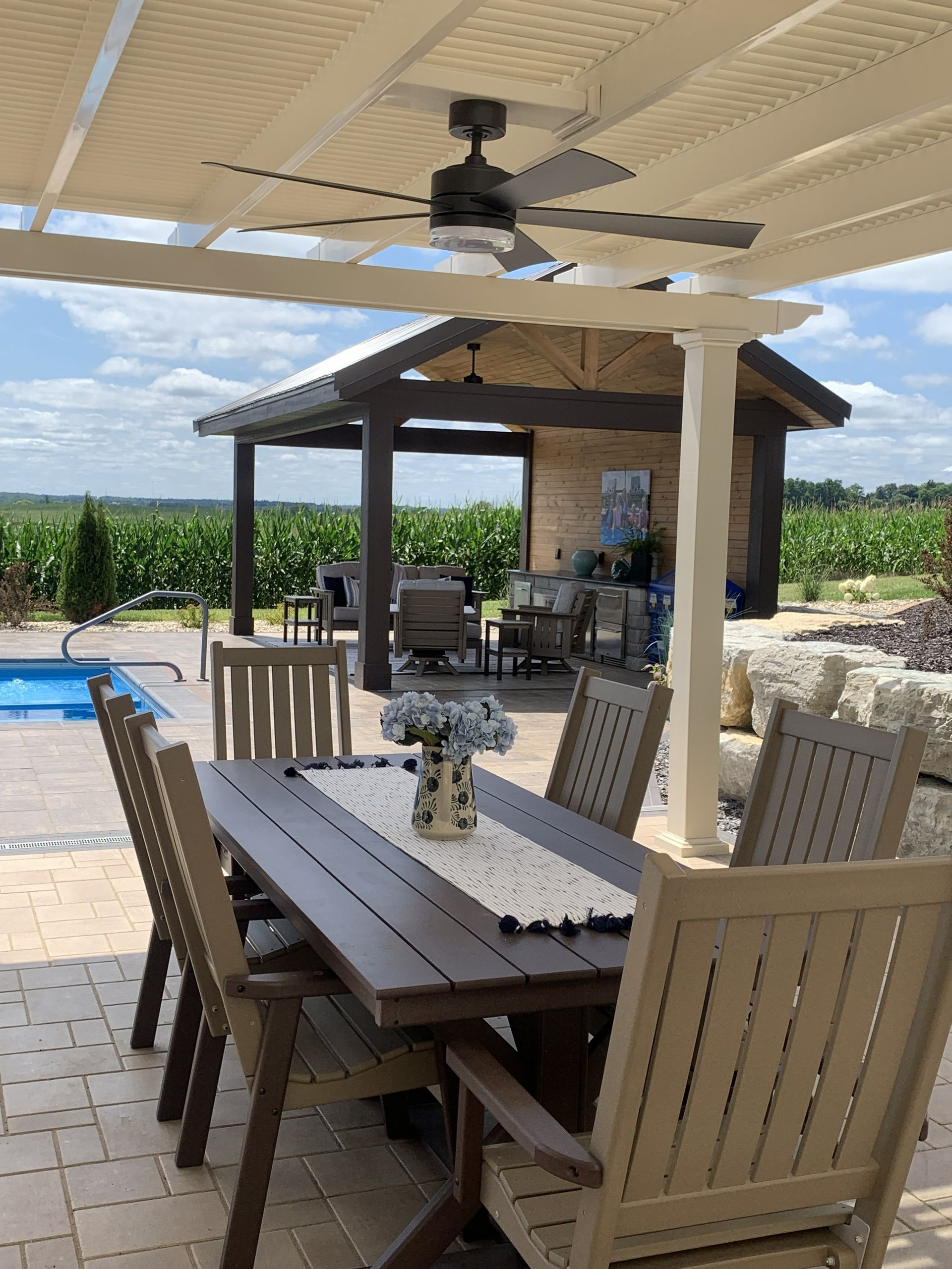 Patio with table, chairs, pergola, pool, and gazebo against a field and blue sky.