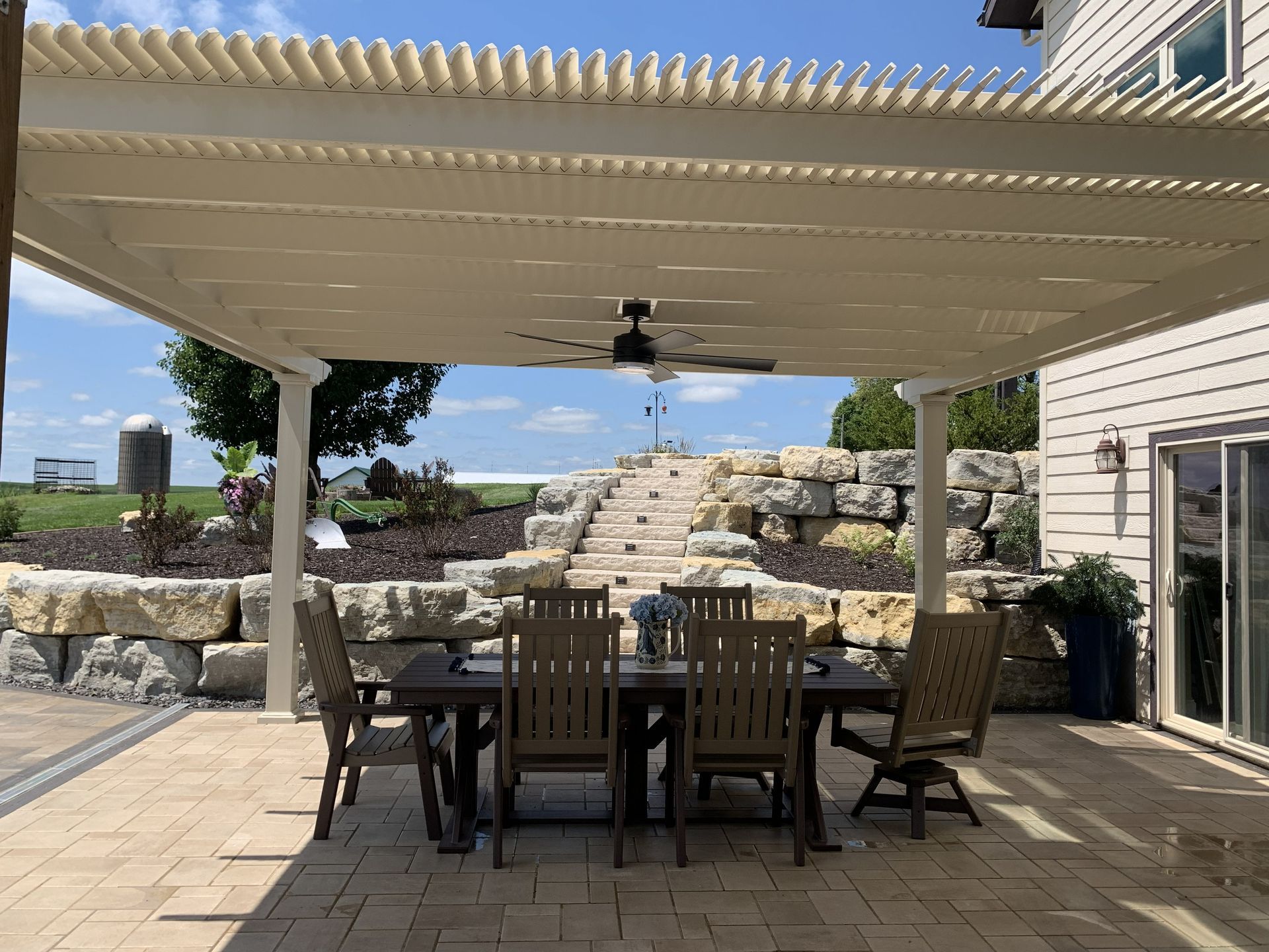 Patio with dining table, chairs, beneath a cream-colored pergola, steps lead to a landscaped area, blue sky.