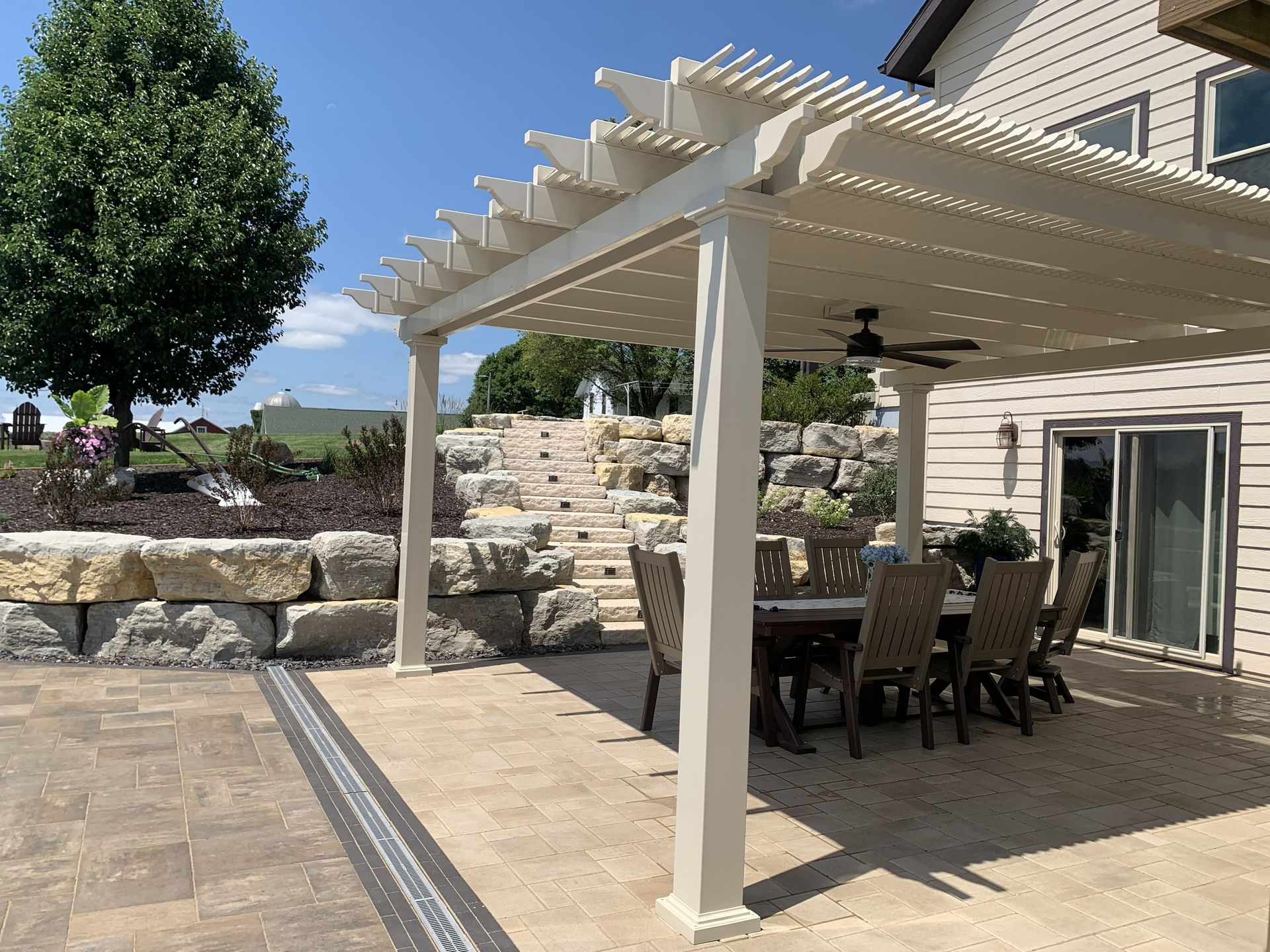 Outdoor patio with dining table under a pergola. Beige structure, stone steps, and blue sky.