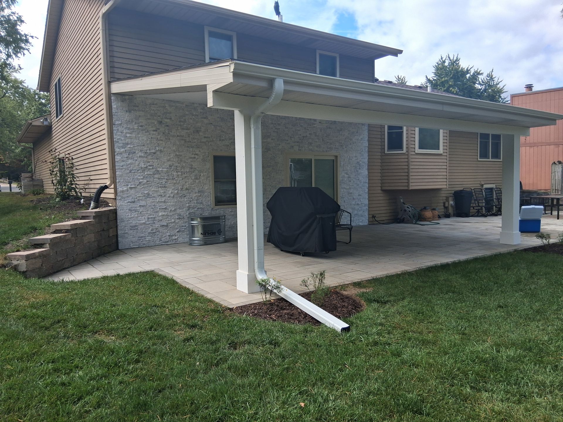 Backyard patio with covered seating area, grill, and two-story house with tan siding.