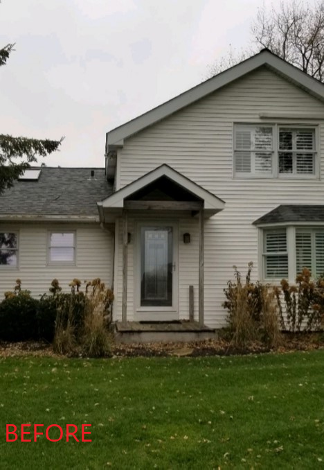 White two-story house with front porch and overgrown landscaping. Overcast sky and green lawn.