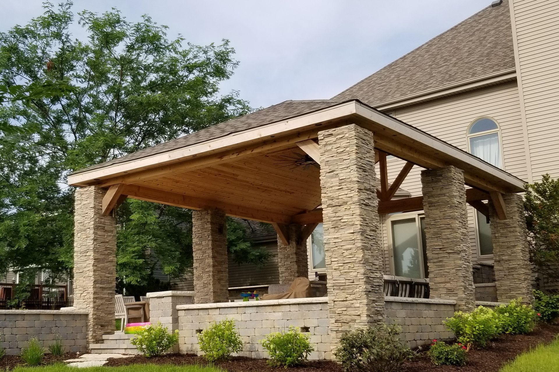 Covered patio with stone pillars and wood ceiling, attached to a brick house.