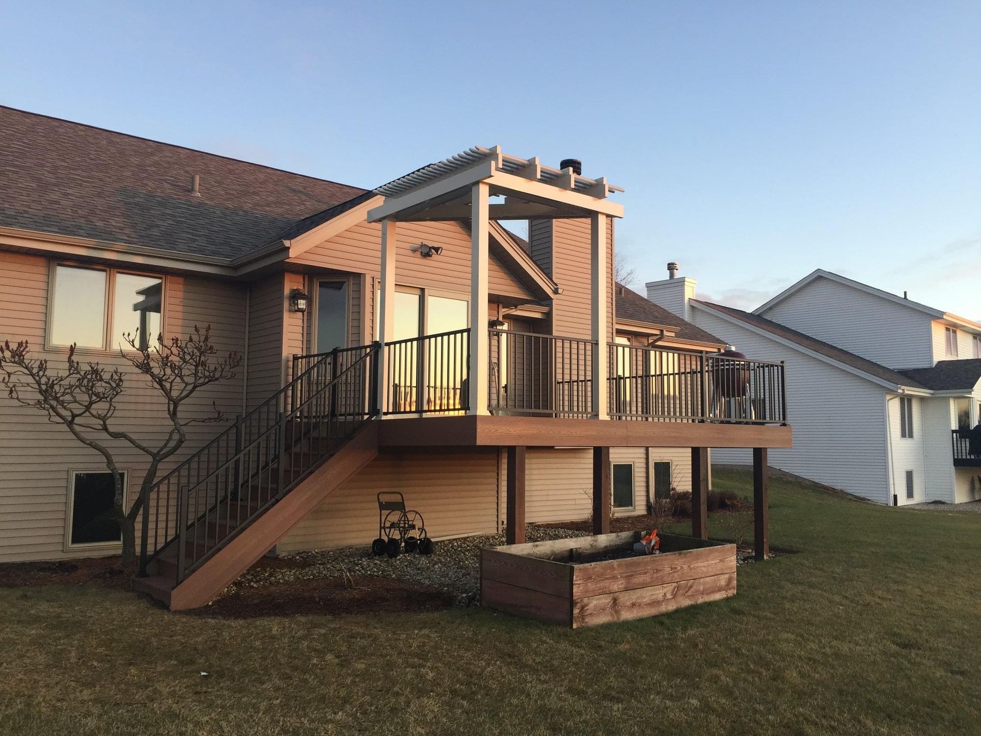 Wooden deck with pergola attached to a two-story beige brick house, overlooking a grassy yard.