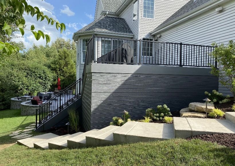 Gray house with a dark gray deck and stairs leading down to a lawn with landscaping.