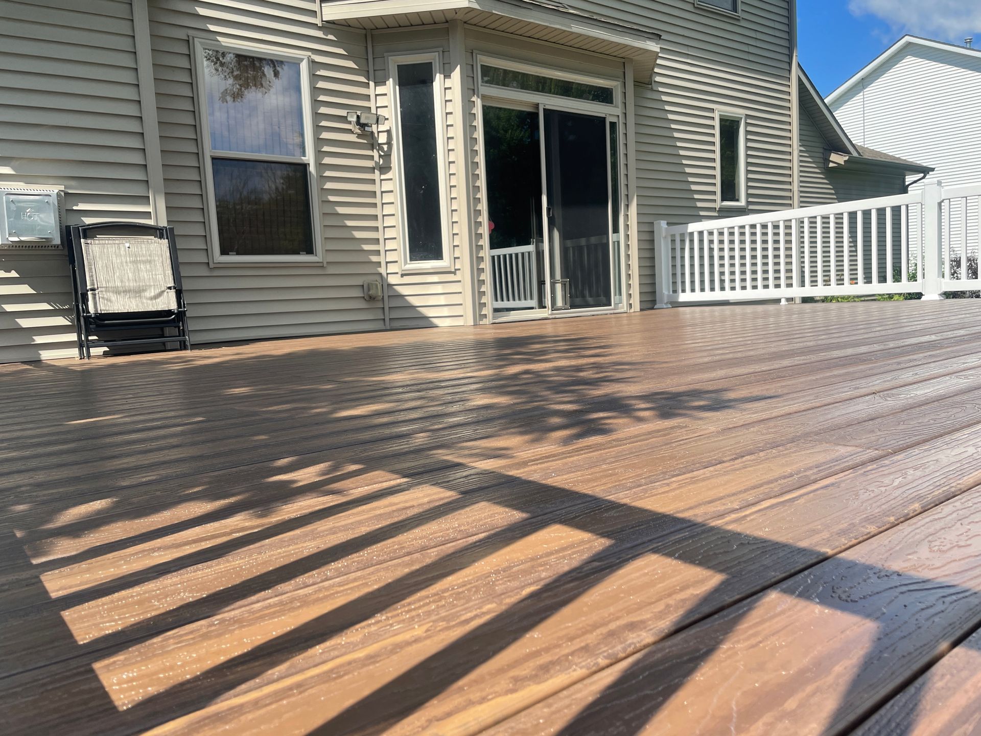 Brown composite deck with shadows, leading to a house with a sliding glass door and white railing.