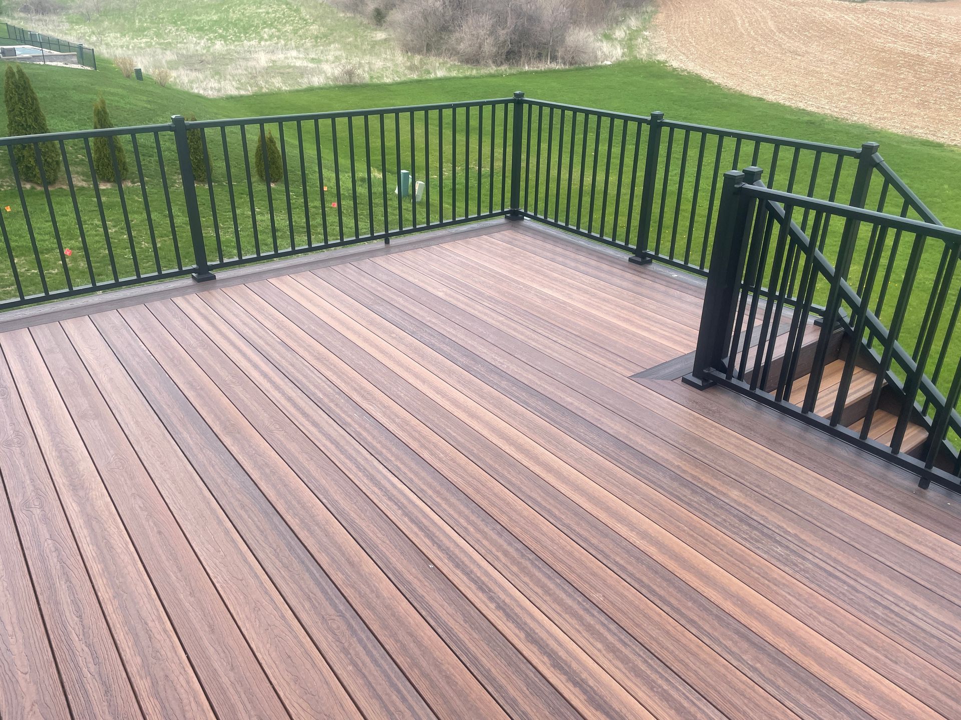 A wooden deck with black railings overlooks a grassy yard and field.
