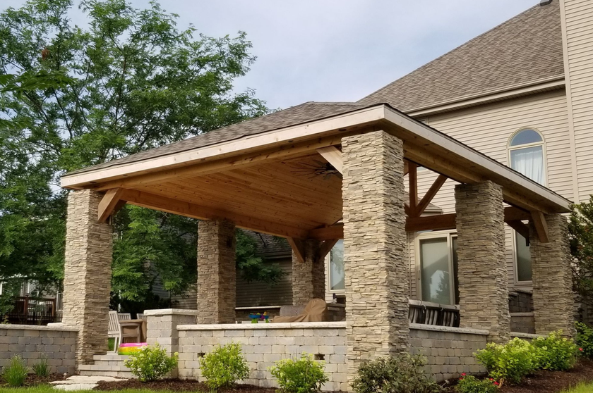 Stone-covered patio with wooden ceiling, adjacent to a brick house. Green plants and foliage are in the background.