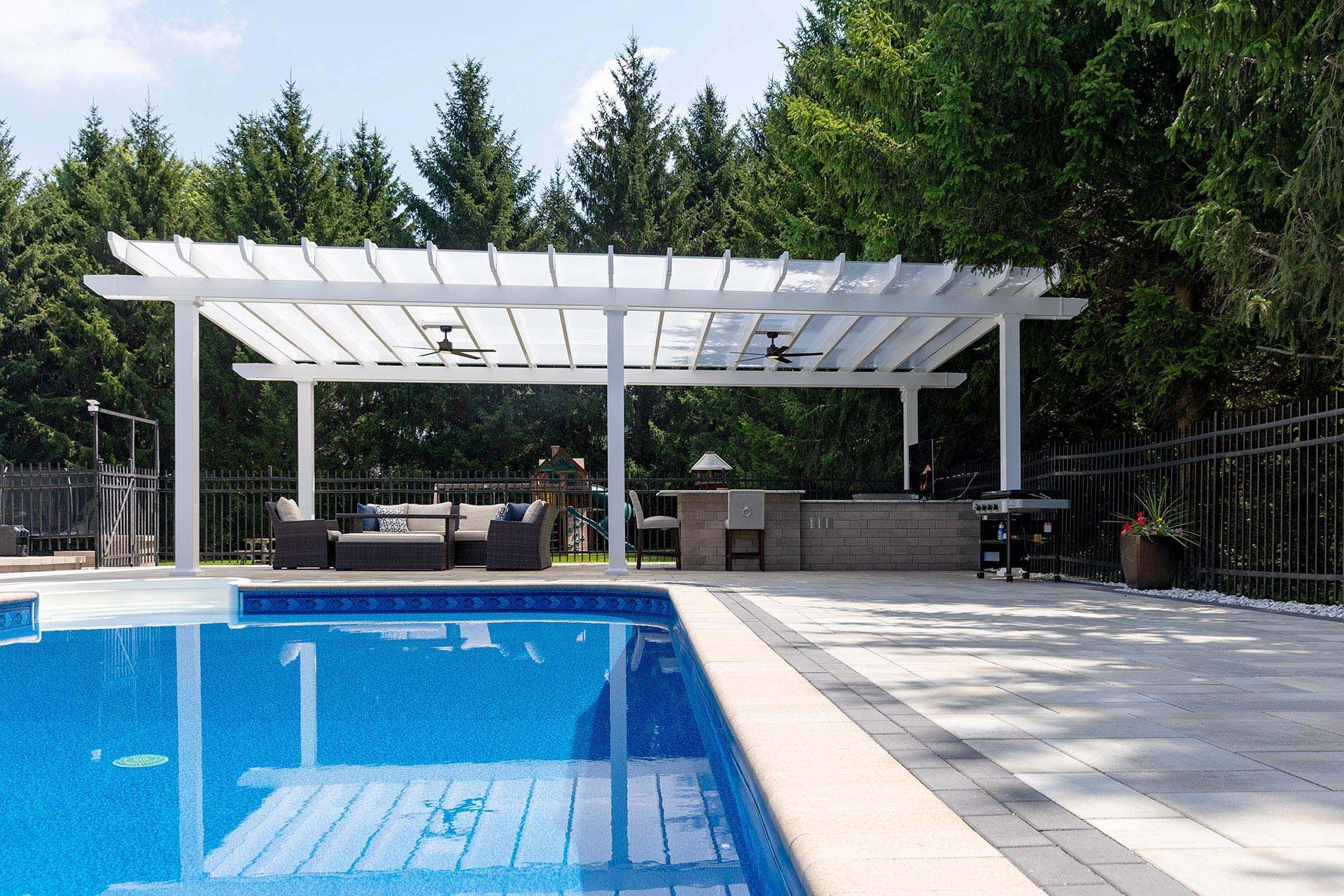 A pool area featuring a white pergola with fans over a patio seating area and outdoor kitchen near trees