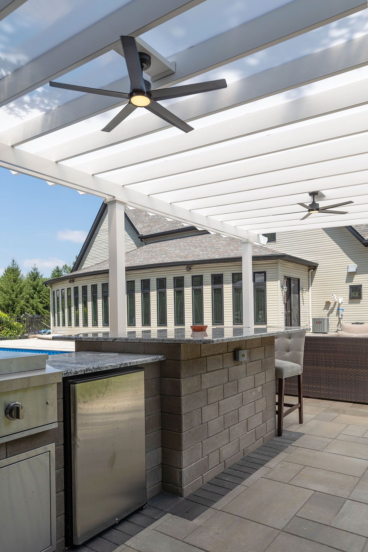 A covered outdoor kitchen with a stone island, stainless steel refrigerator, and ceiling fans on a paved patio