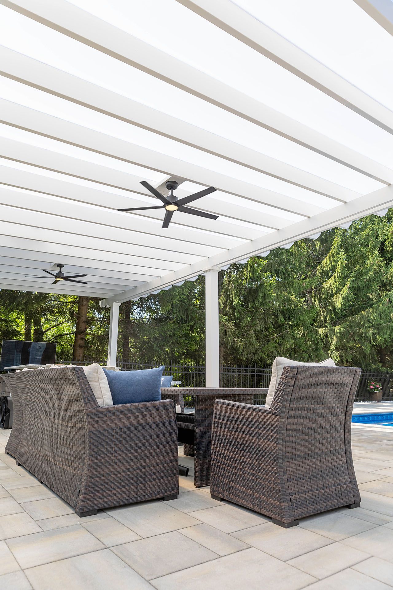 A patio furniture under a white louvered pergola with ceiling fans by a pool
