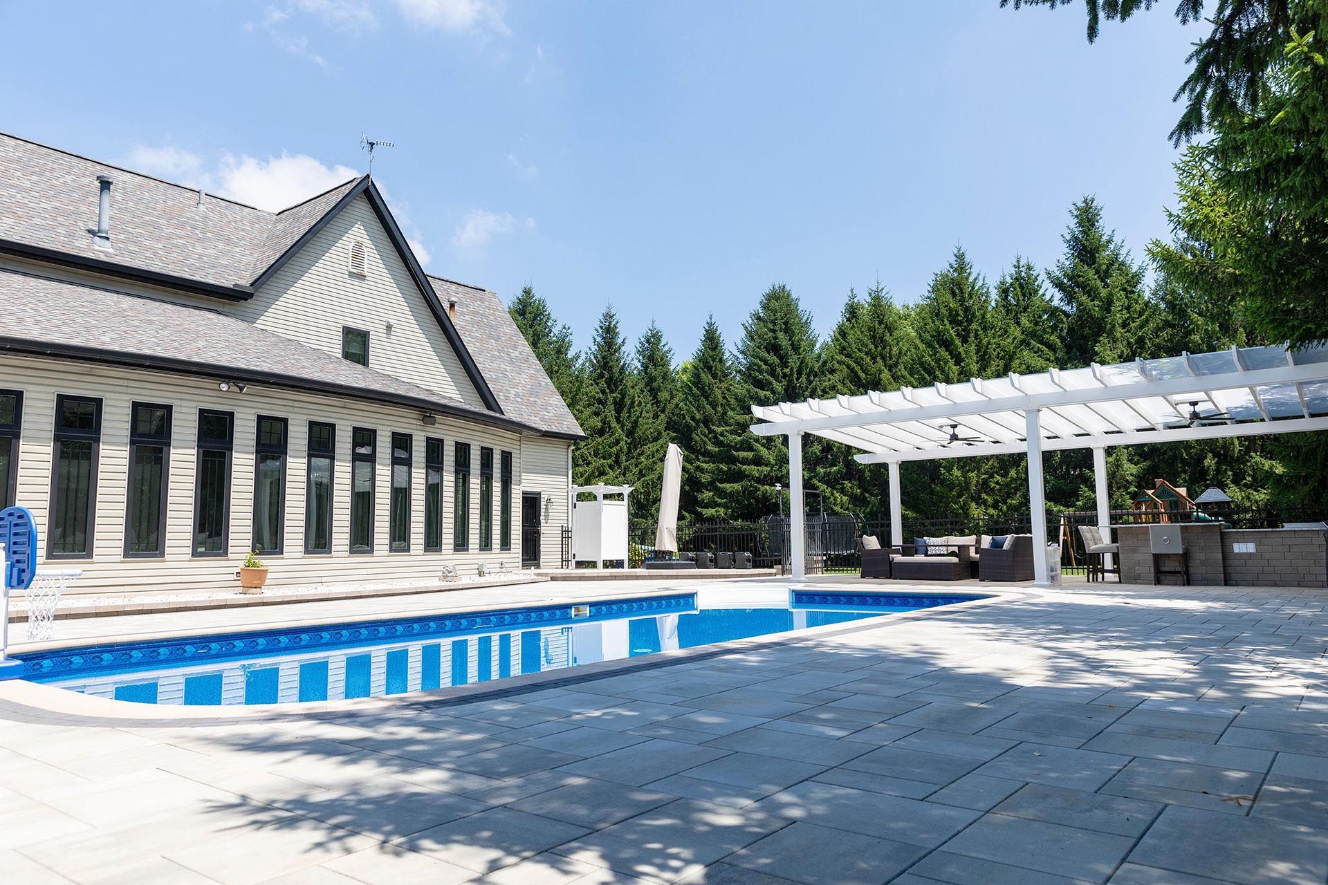A backyard swimming pool with a stone patio, next to a house and a white pergola