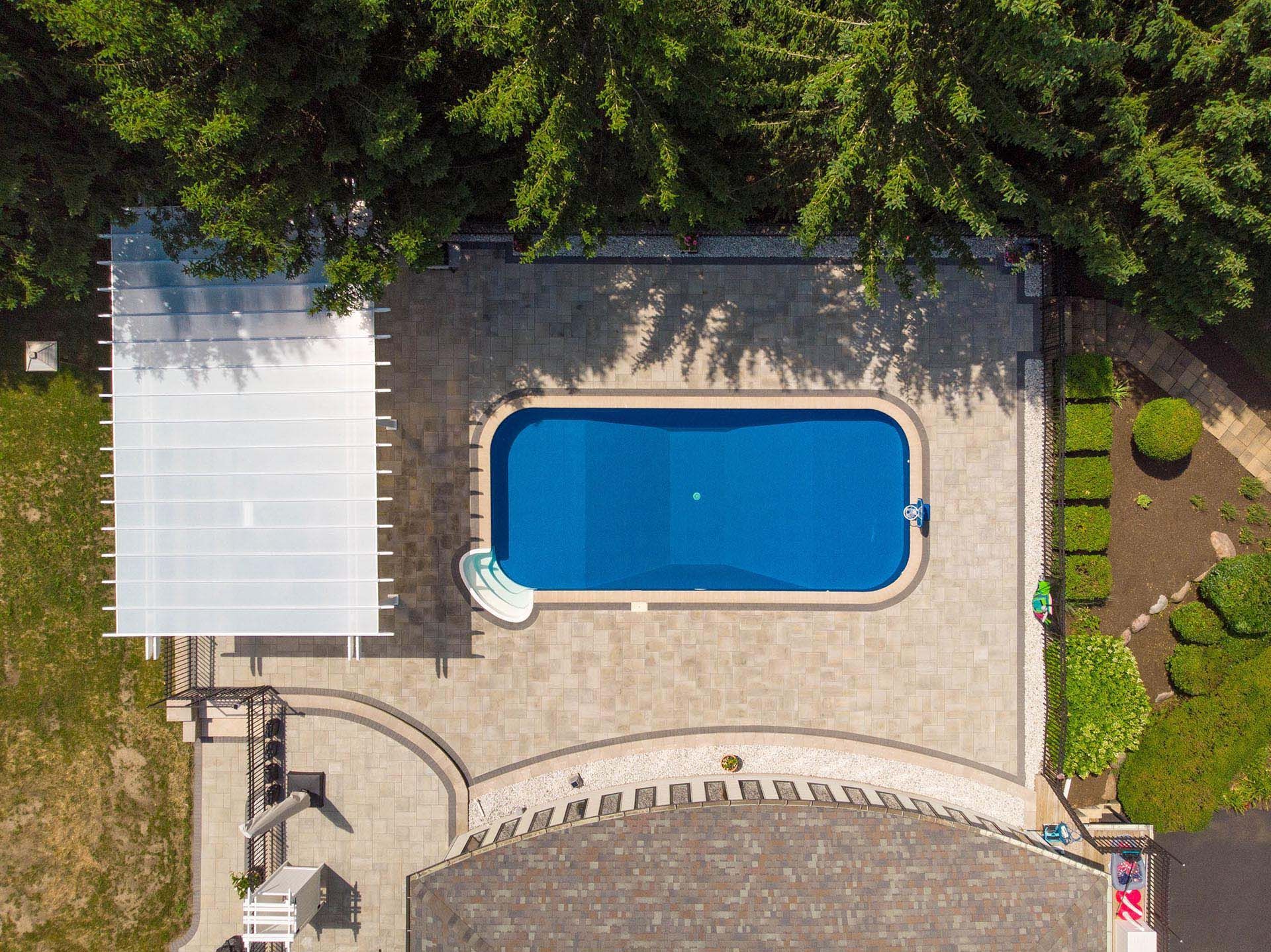 A bright blue rectangular swimming pool sits in a paved patio surrounded by trees and a shaded pergola