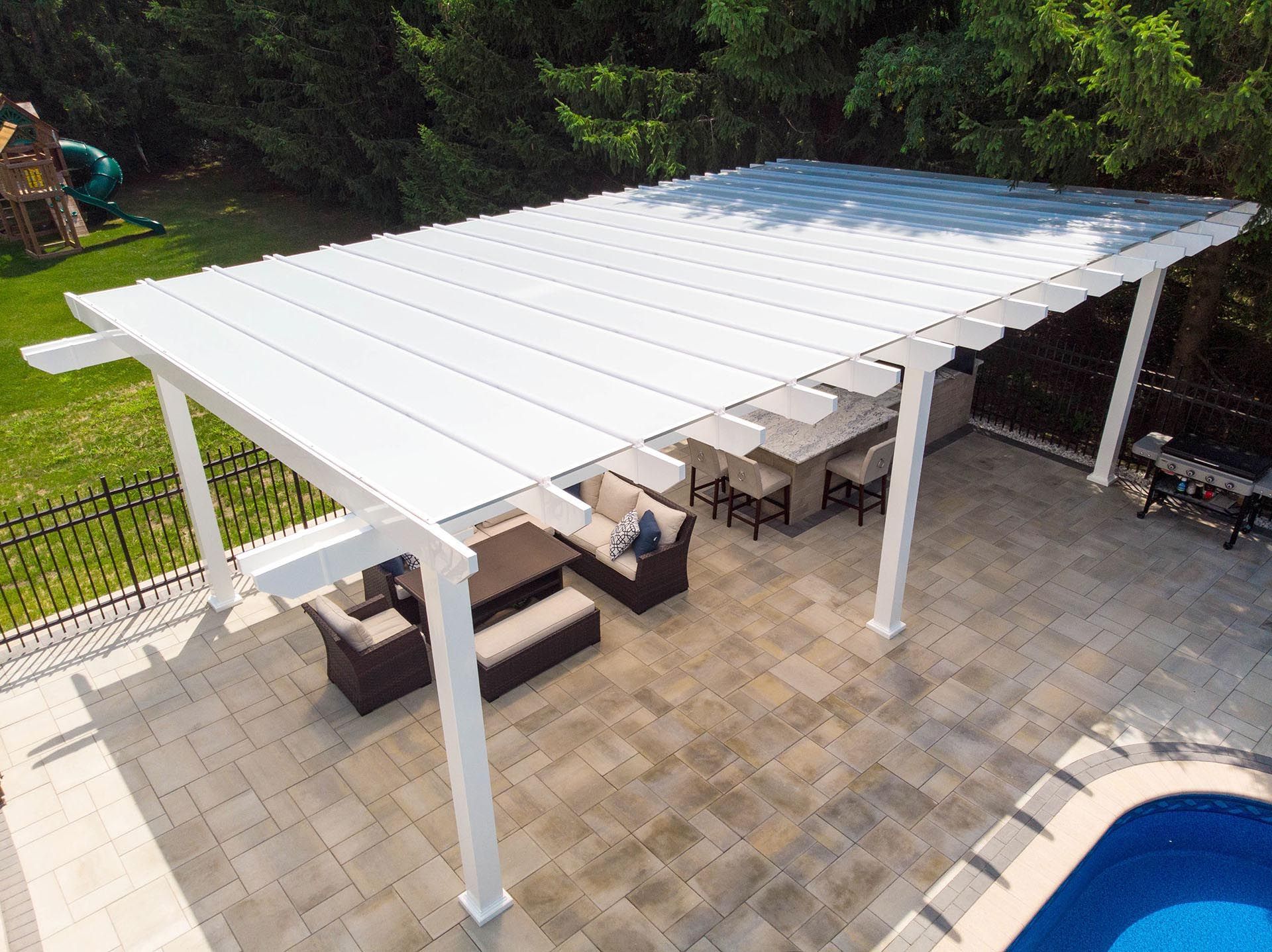 High-angle view of a white pergola covering a stone patio with outdoor furniture, adjacent to a pool and grassy yard