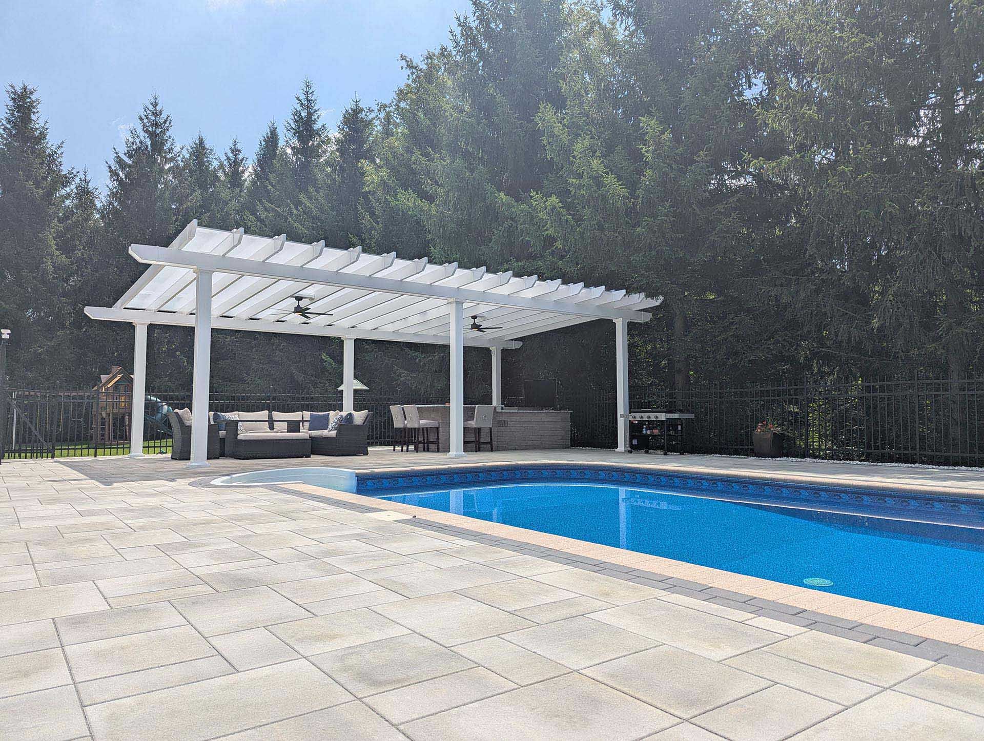 A white pergola stands on a paved patio beside a swimming pool