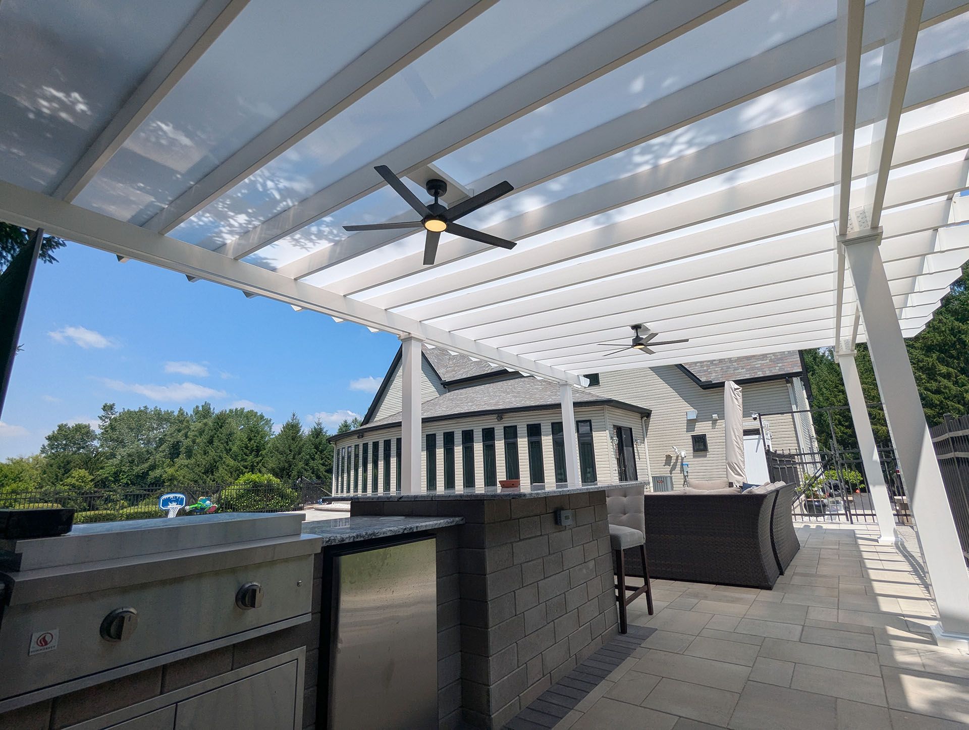 An outdoor kitchen and patio under a white pergola, featuring a stainless steel grill, stone counter, and ceiling fans
