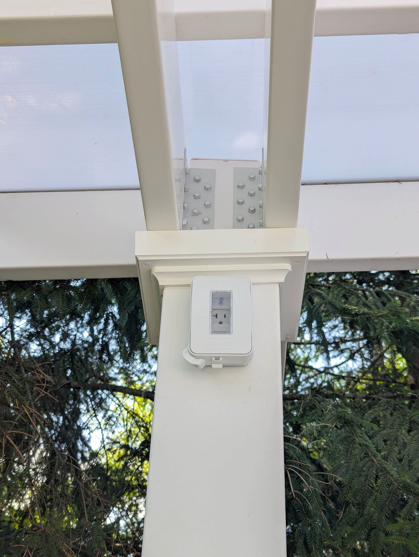 A white electrical outlet with a protective cover mounted on a white pergola post
