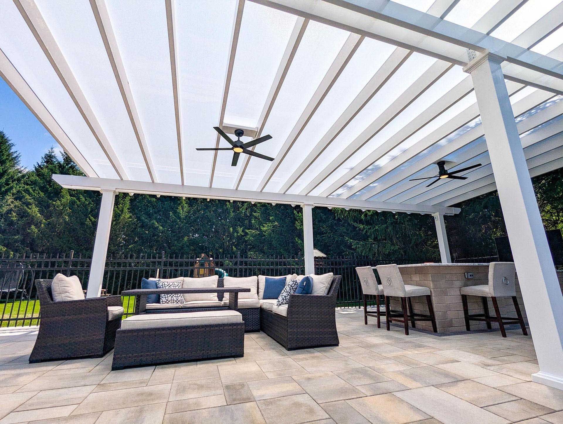 A white pergola covers a patio with dark wicker outdoor furniture, ceiling fans, and a stone bar with stools