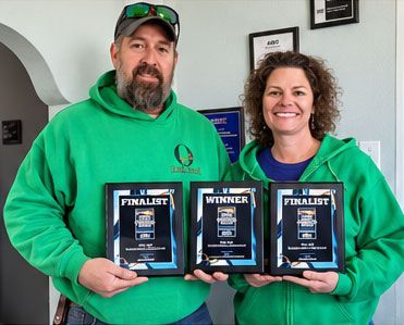 Two people in green hoodies hold three framed award plaques and smile indoors.