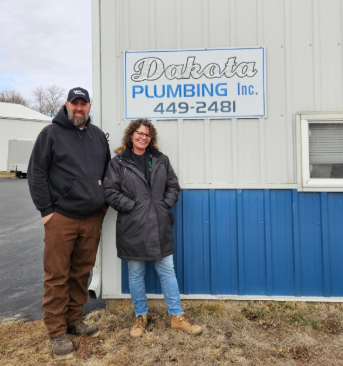 Two people standing beside a Dakota Plumbing Inc. sign on a building exterior