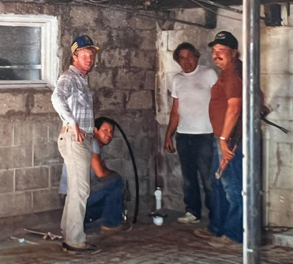 Four workers stand in an unfinished cinder block room near plumbing and tools.