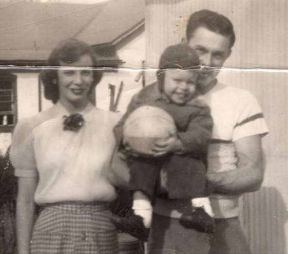 Three people standing outdoors, one holding a child, in front of a house