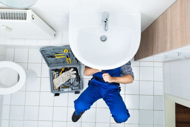 Plumber in blue uniform working under a bathroom sink beside tools and a toilet, top-down view