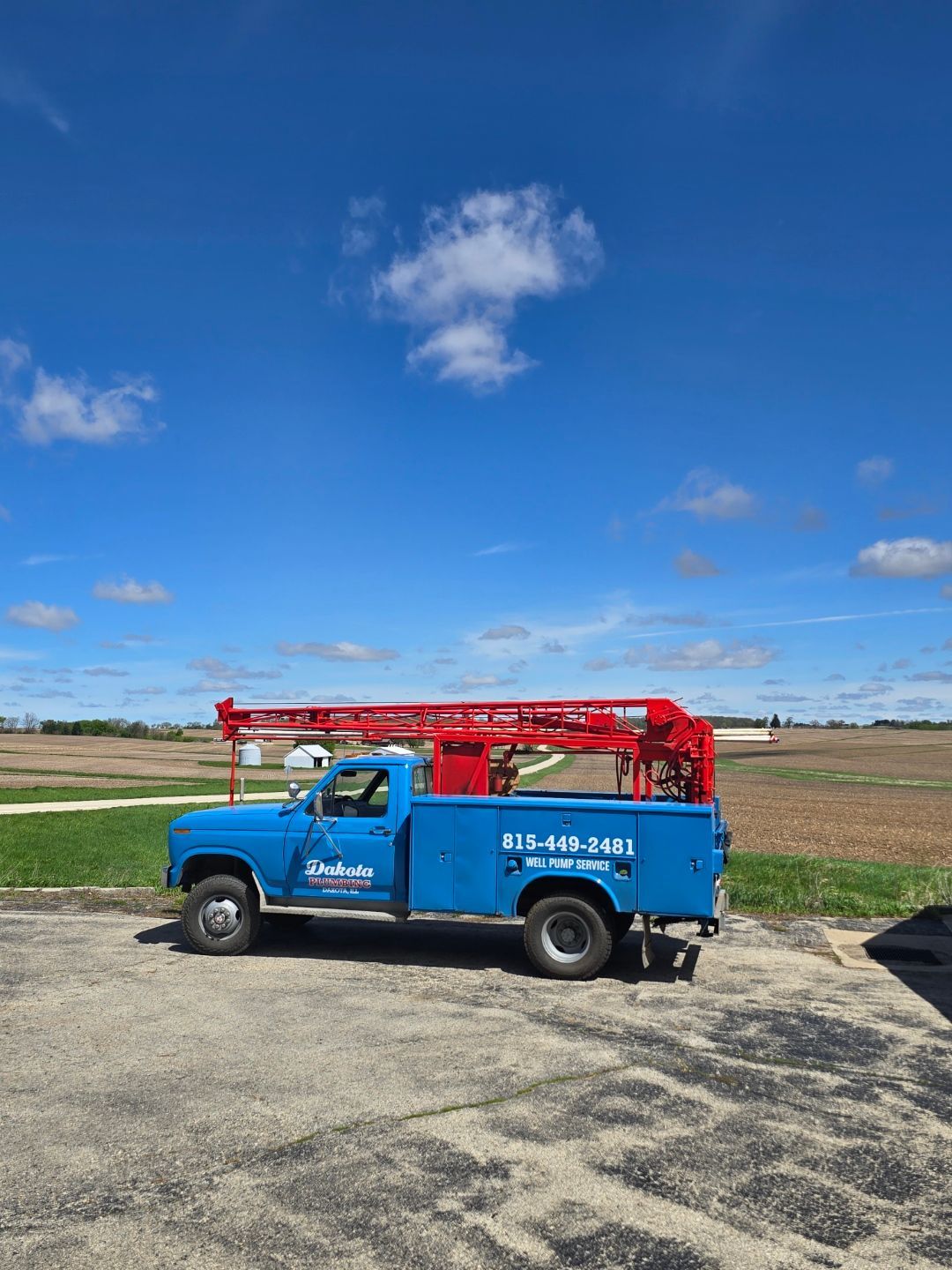 Blue utility truck with red ladder parked on a gravel road in an open field under a blue sky.