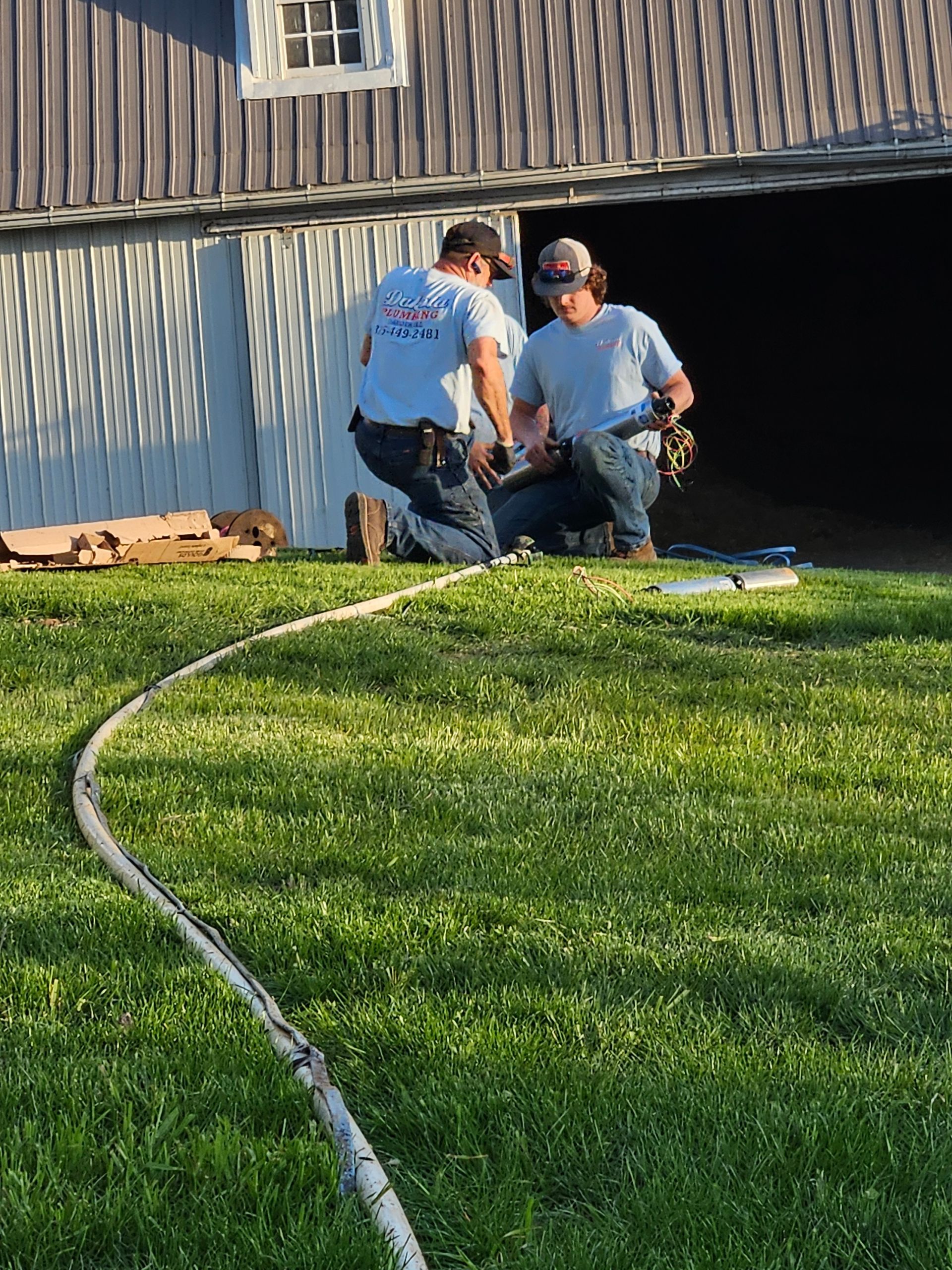 Two workers kneel by a house wall, inspecting equipment beside a grassy yard and hose.