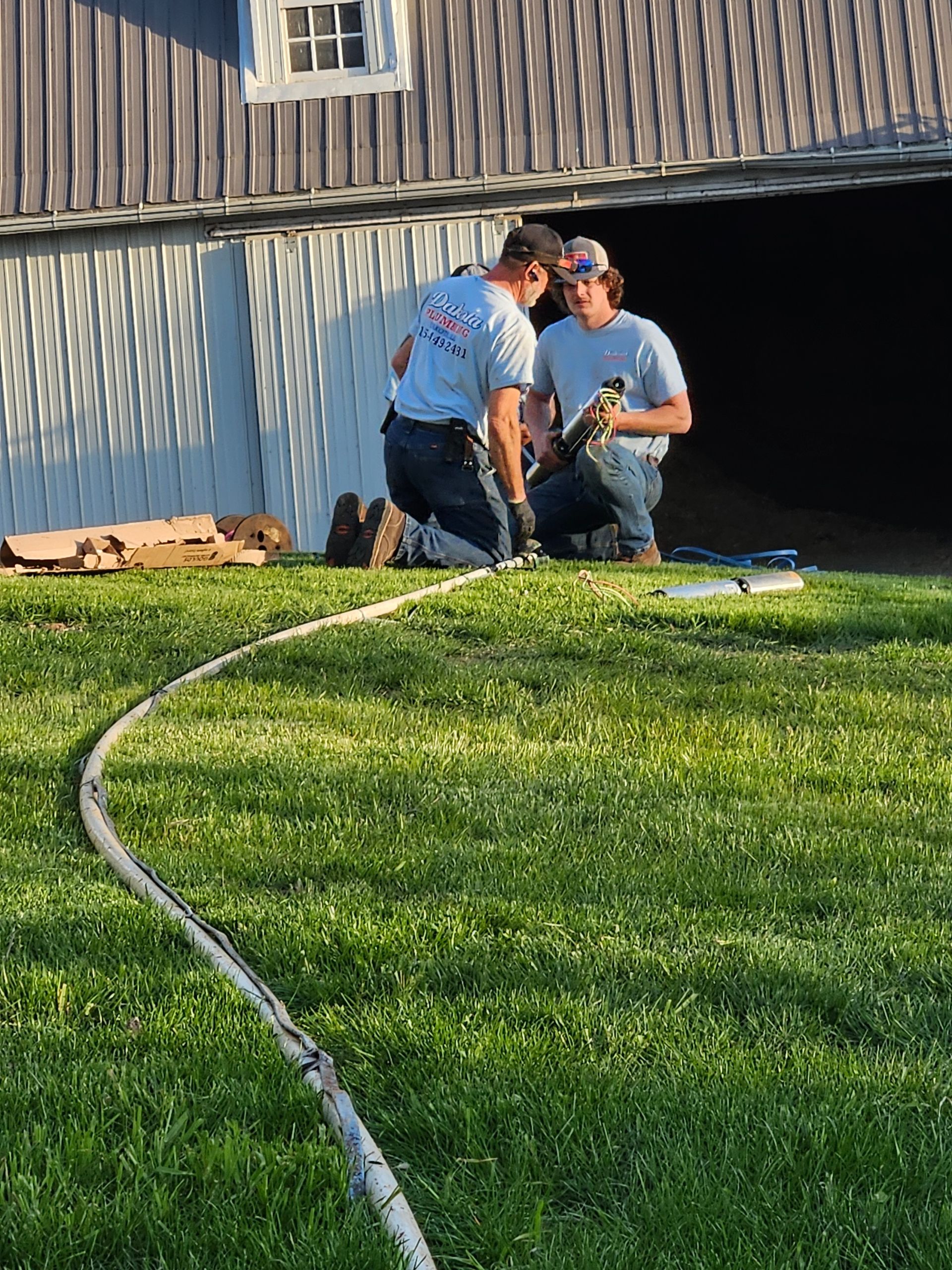 Two utility workers kneel on grass near a building, inspecting equipment beside a dark open doorway.
