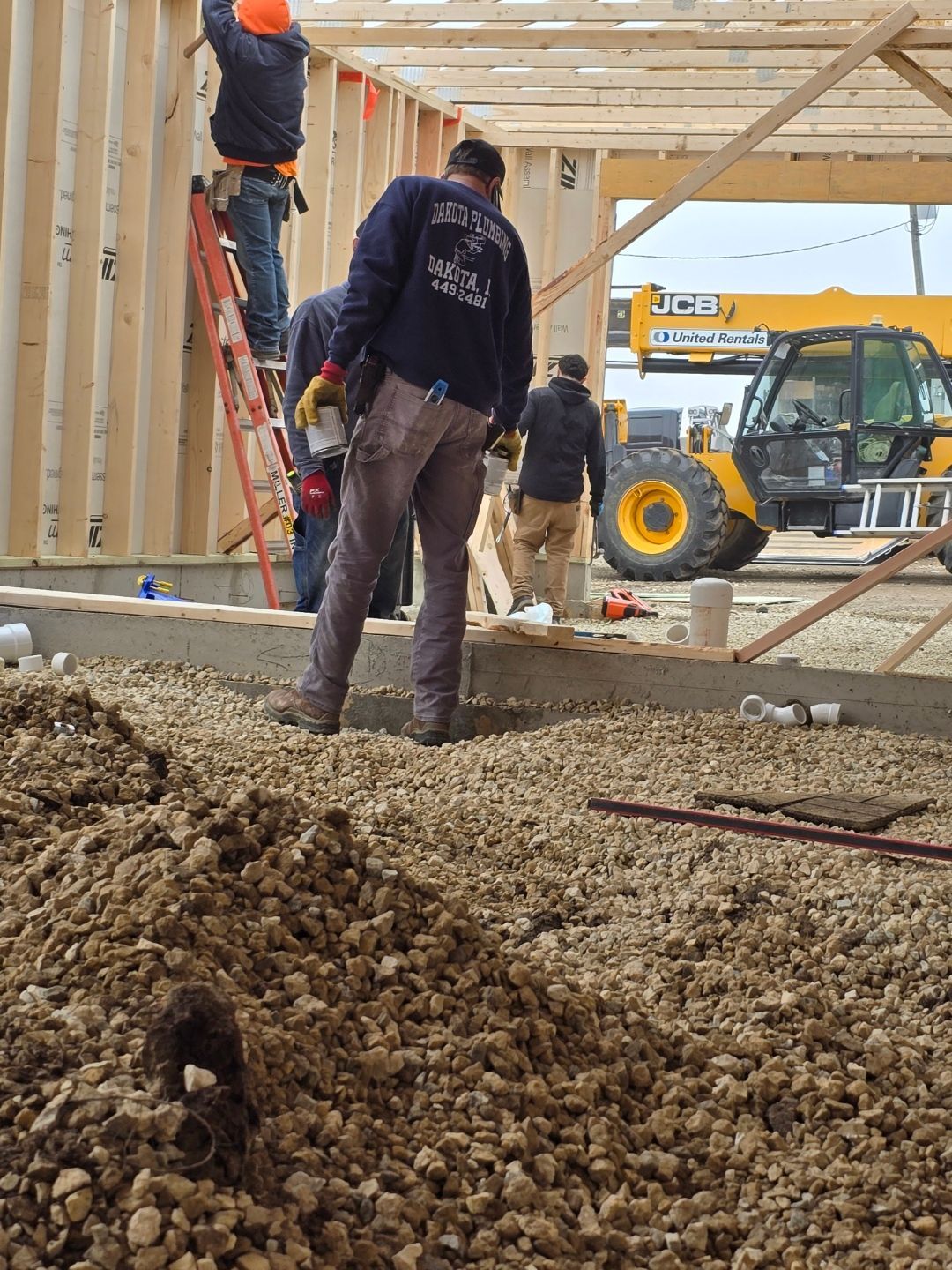 Construction workers framing a wooden building foundation, with a yellow loader in the background.