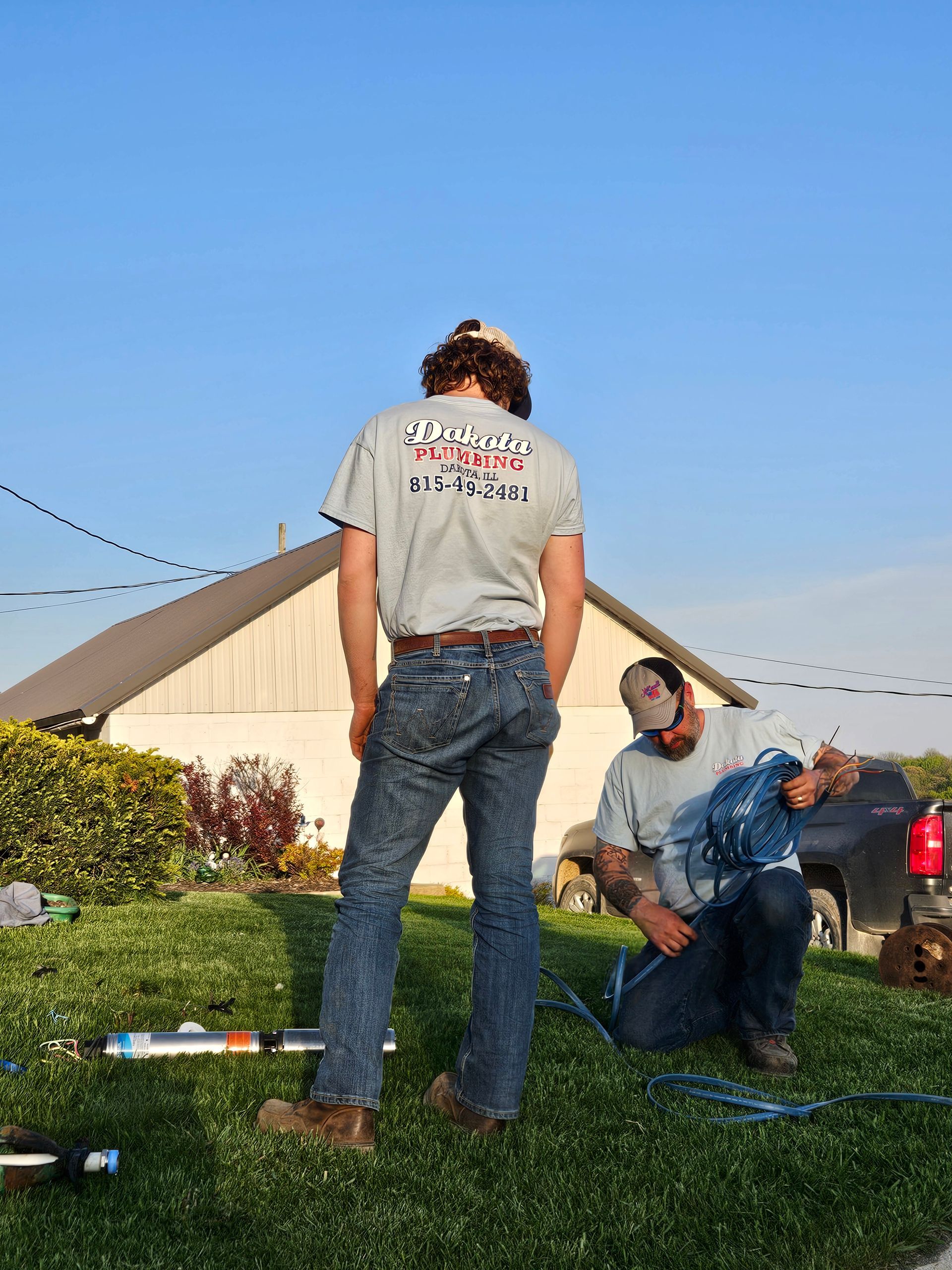 Three people on grass near a tent, one standing and two sitting, under a blue sky.