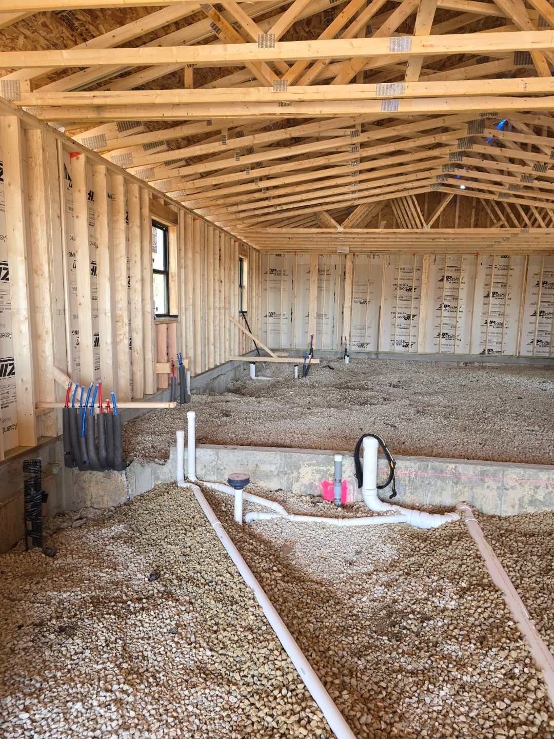 Framed house interior under construction with exposed wood trusses, gravel floor, and plumbing pipes.