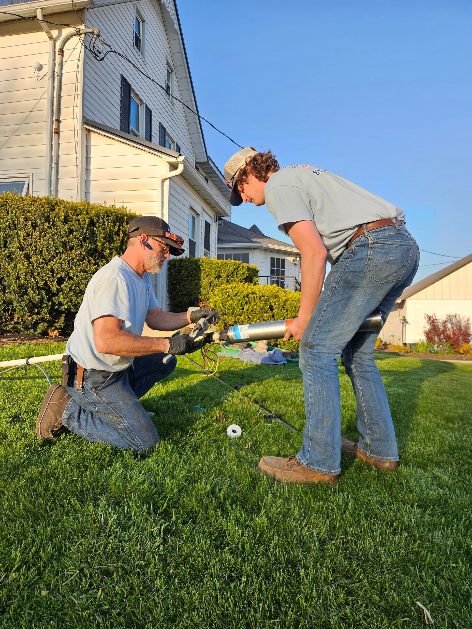 Two people kneel and bend on a lawn, working together on a garden hose near a house.
