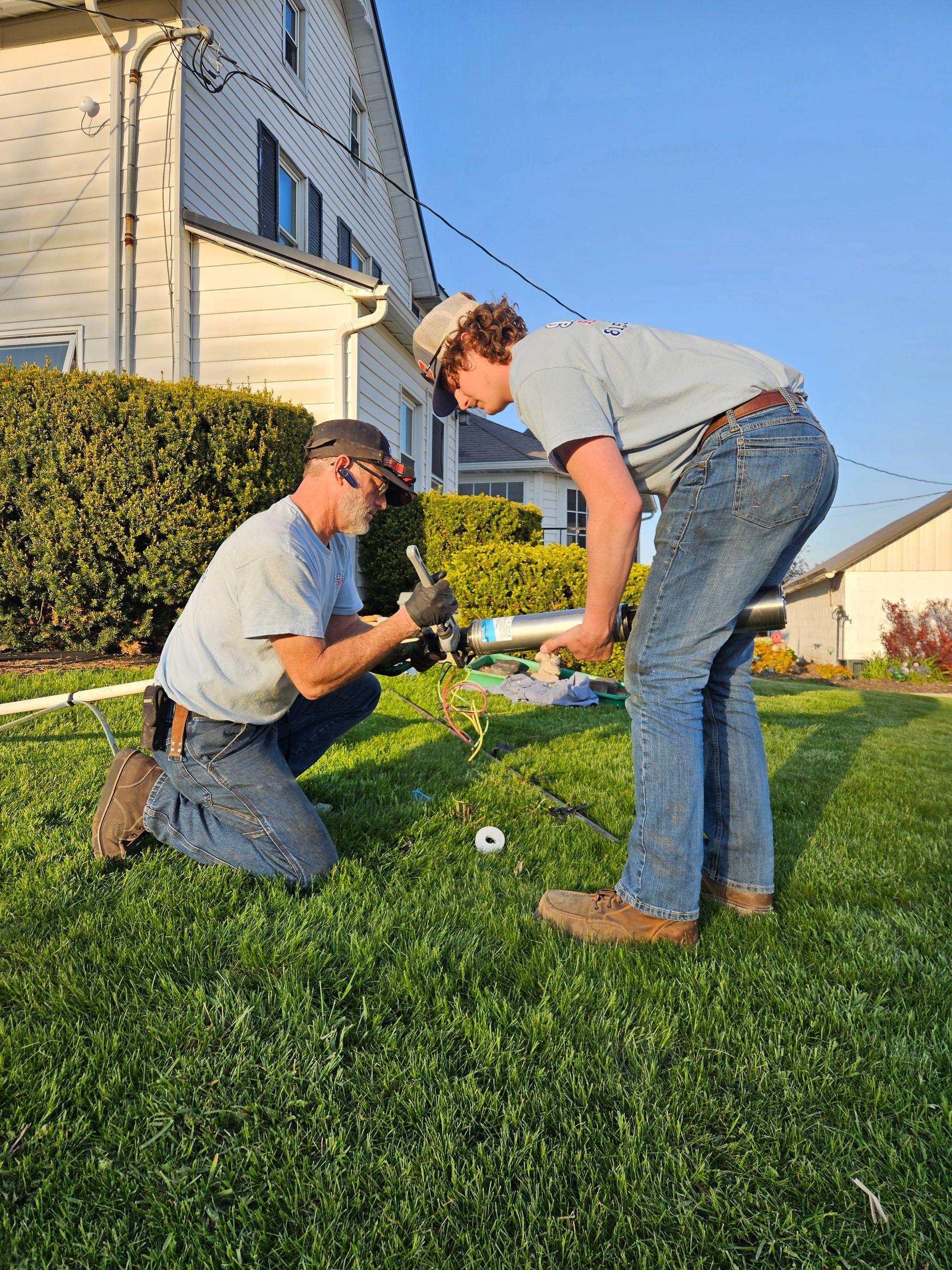 Two people kneeling on a lawn, working on a small garden near a house in evening light.