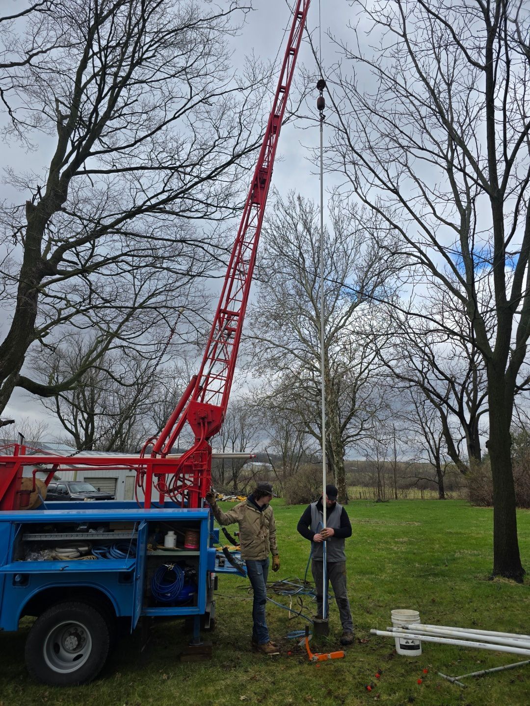 Two workers beside a red boom lift in a park with leafless trees, inspecting equipment on the ground.