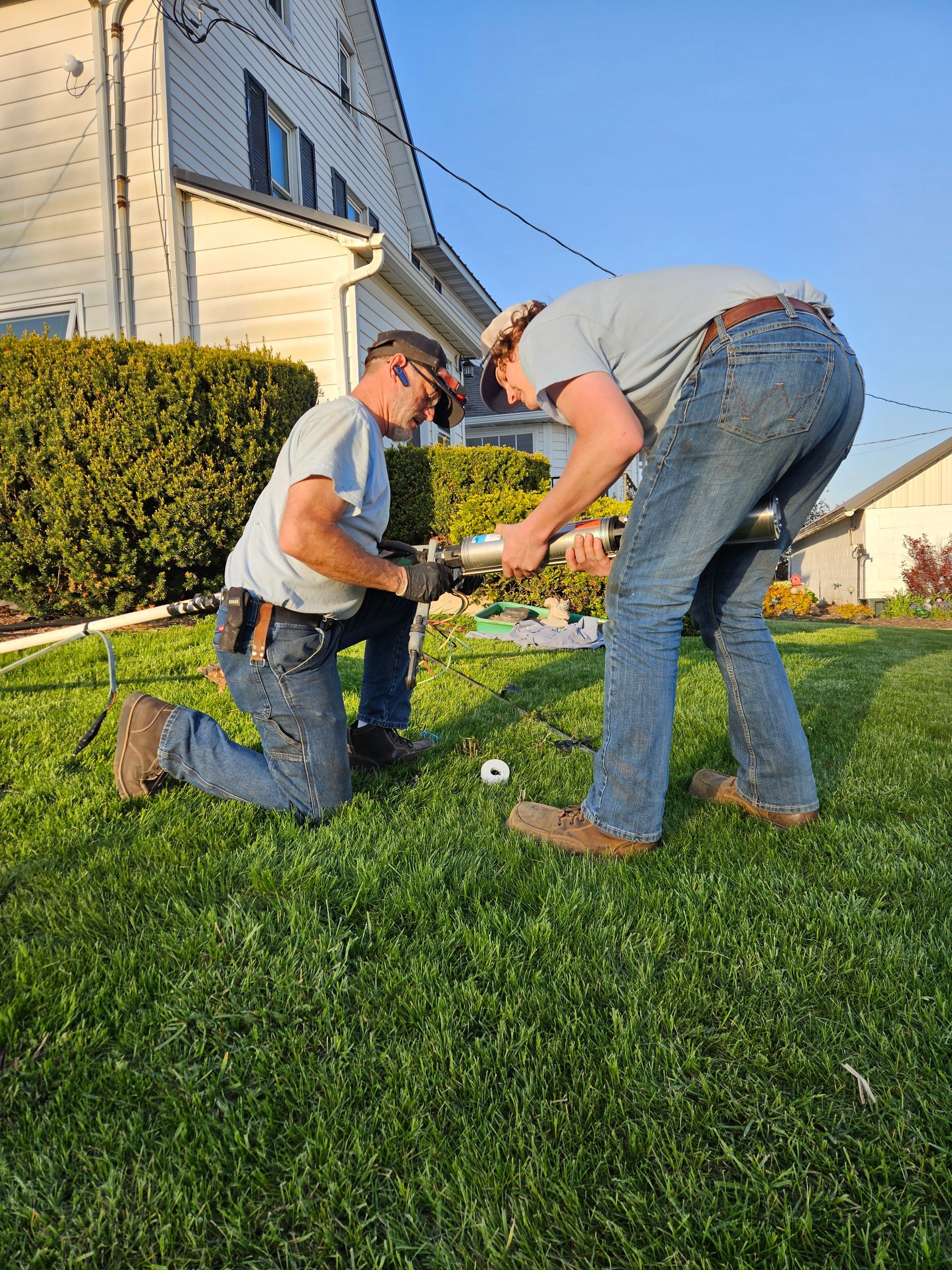 Two people kneel in a yard, measuring and working on the grass beside a house in sunlight.