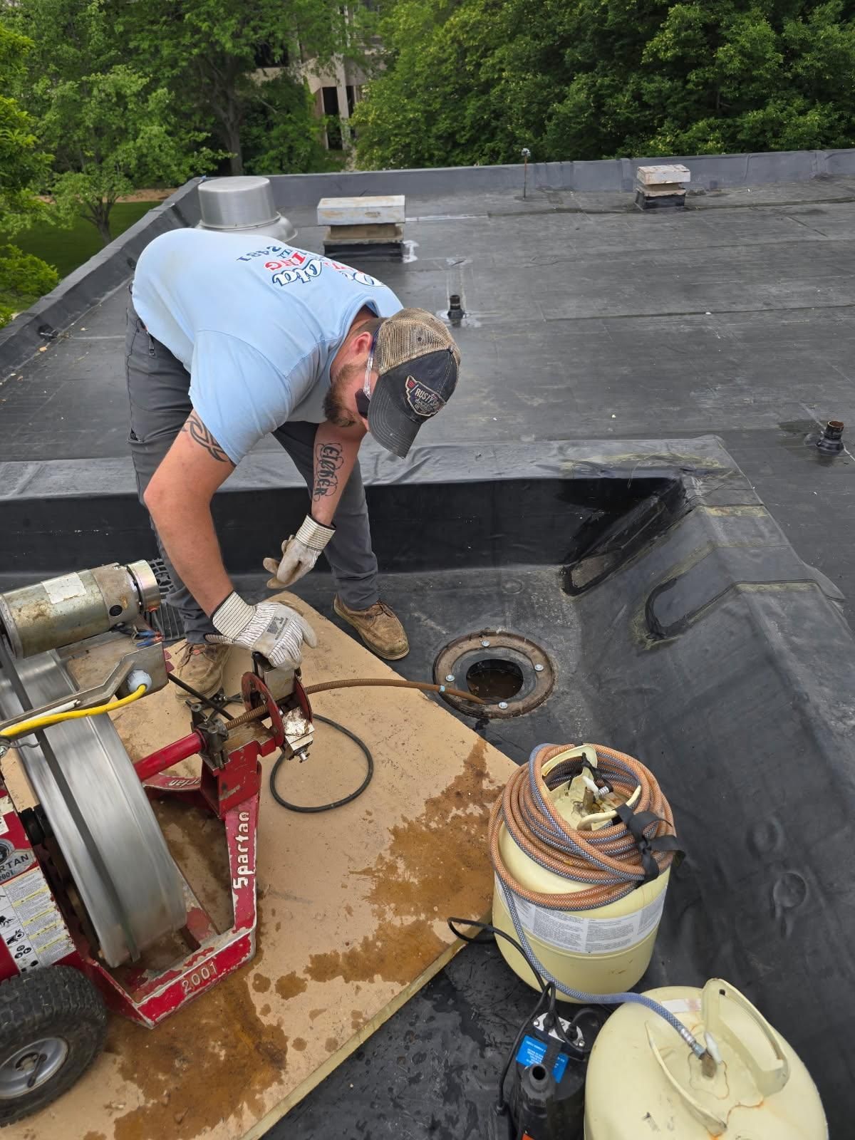 Worker cleaning debris from a rooftop drain beside hoses and equipment.