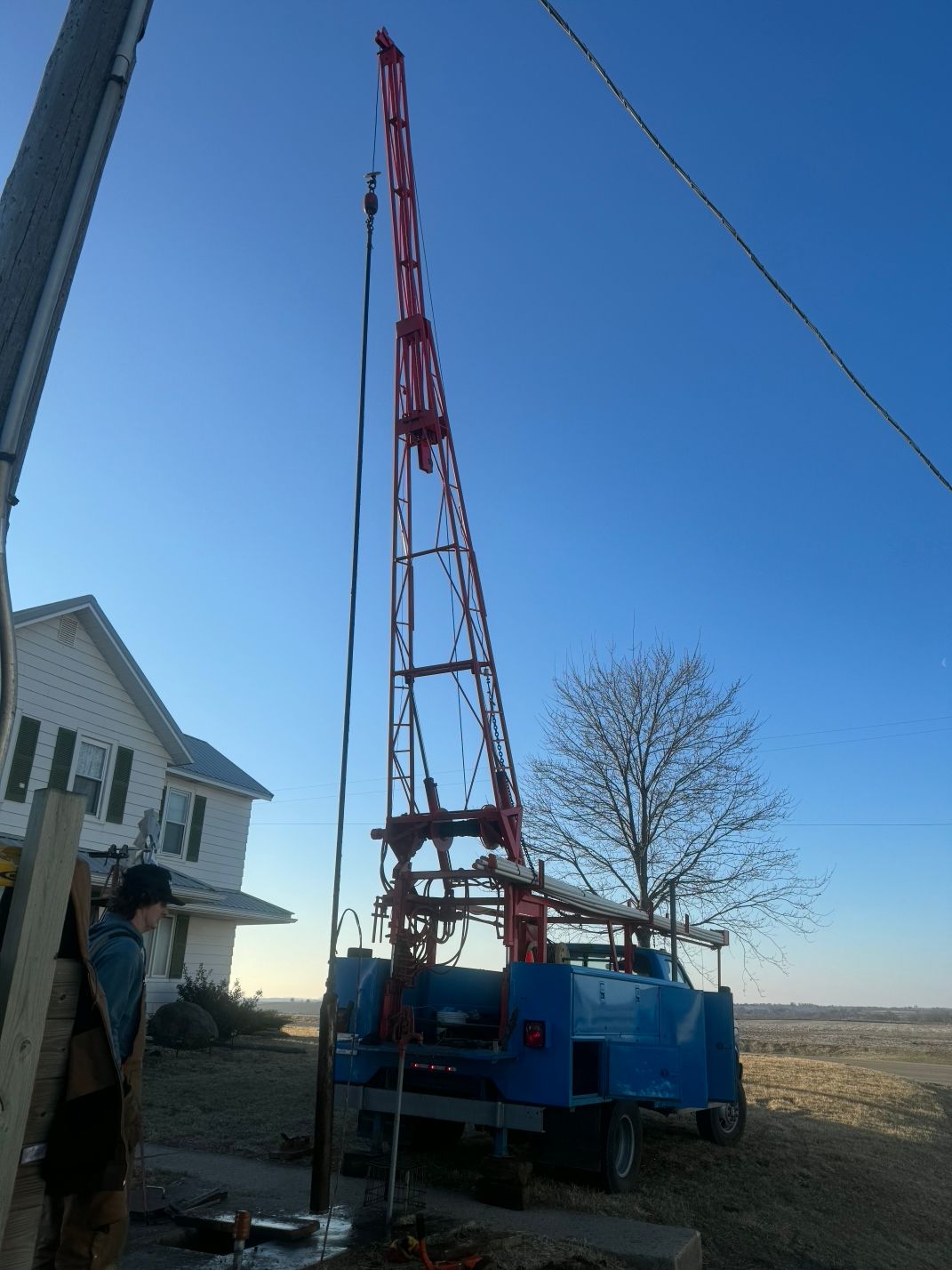 Blue drilling rig beside a house in a rural field under a clear sky