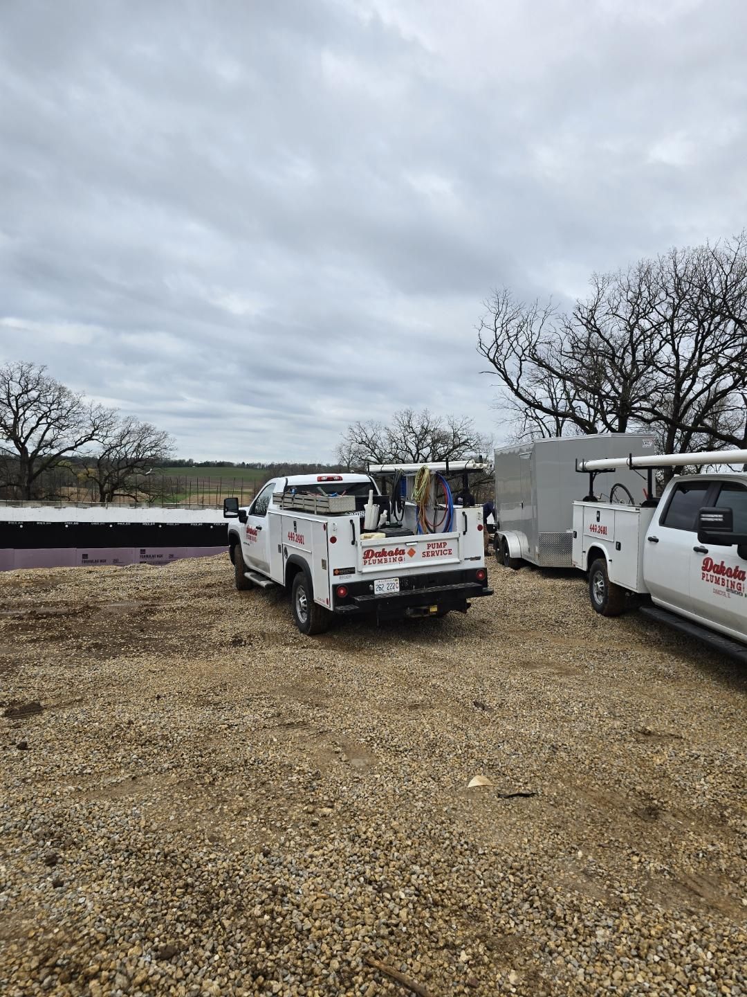 Two white utility trucks parked on gravel under a cloudy sky, with a flat field in the background