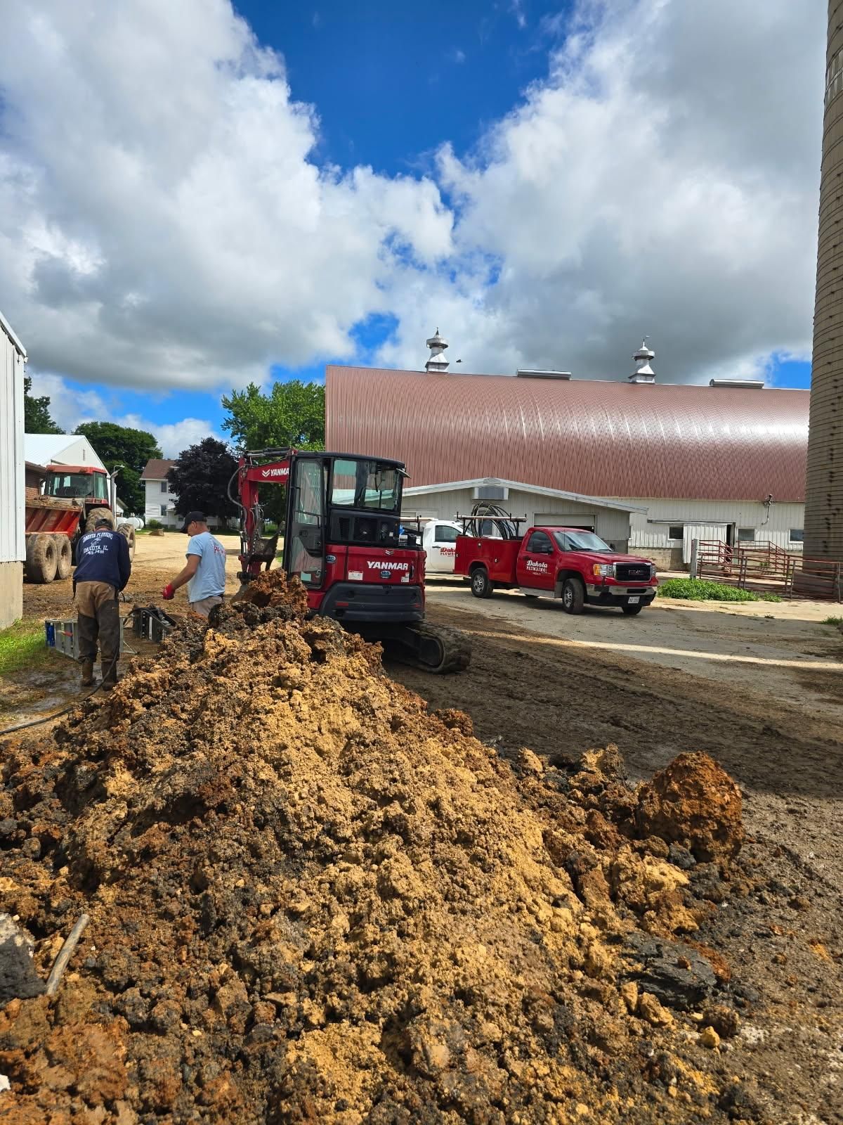 Construction site with dirt pile, excavator, and red trucks beside a large building under cloudy sky