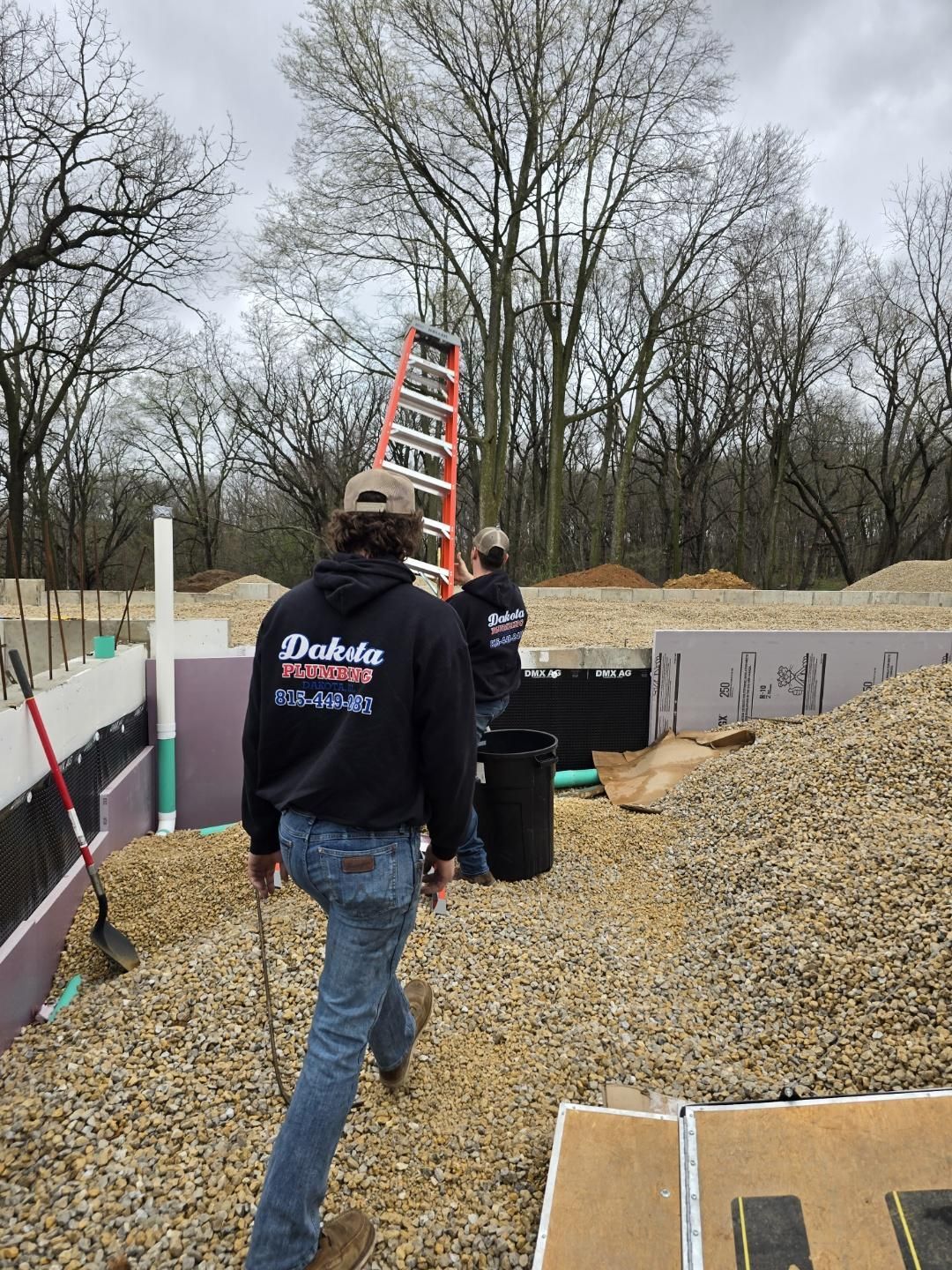 Workers carrying a ladder at a construction site with gravel and partially built foundation walls.
