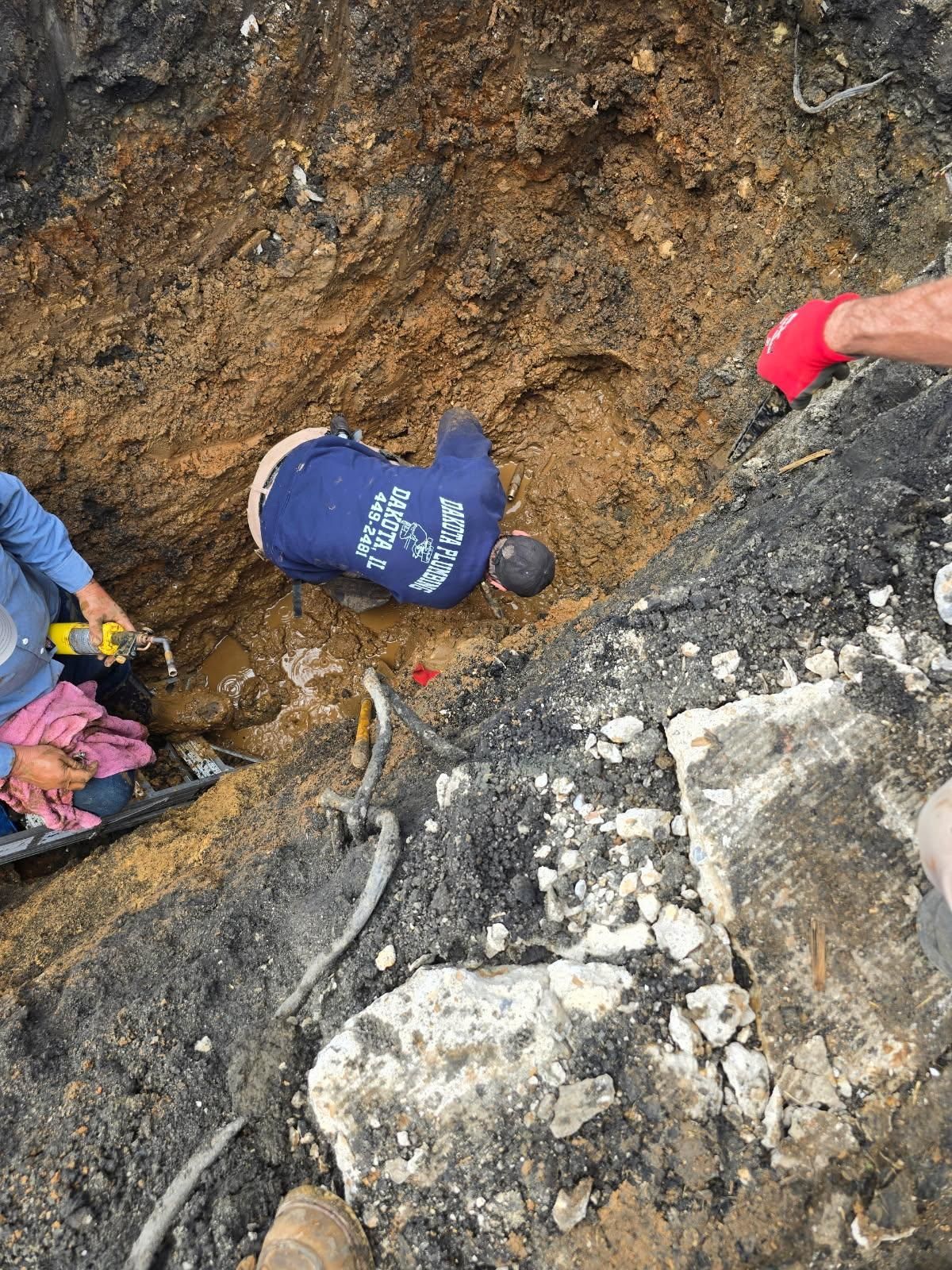 People working in a muddy excavation pit beside broken concrete and exposed roots