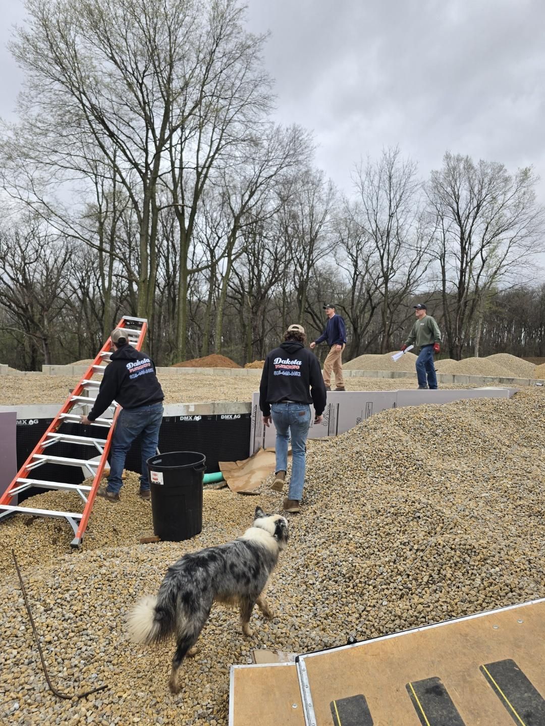People on a gravel mound at a construction site, one carrying a ladder, with a dog in the foreground.