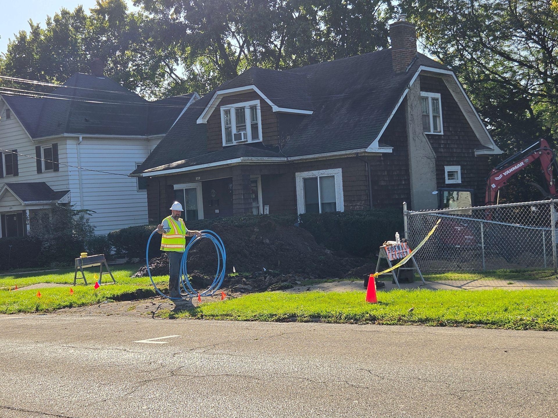 Worker in neon vest on a suburban lawn, with orange cones and hose in front of a dark house.