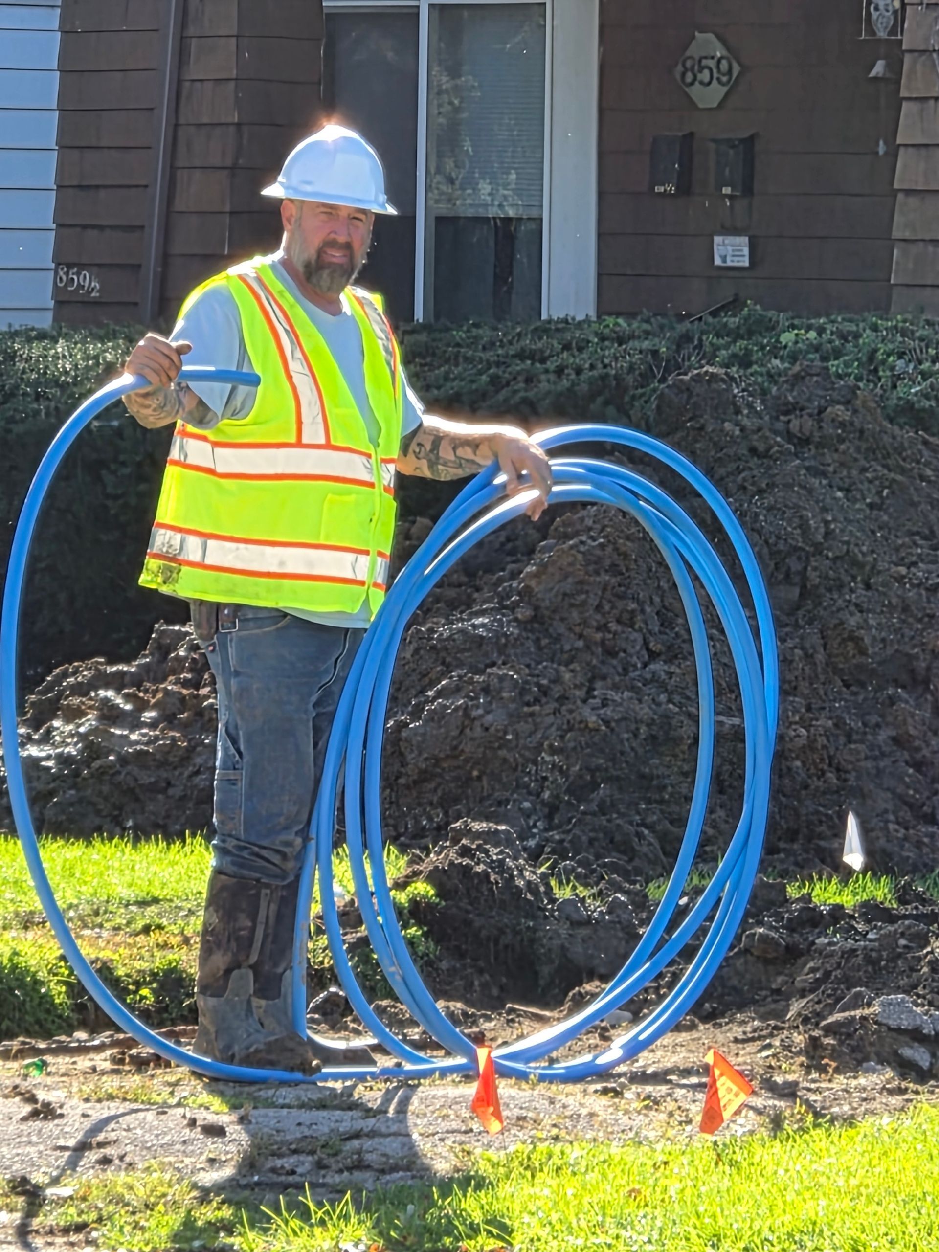 Worker in hard hat and safety vest holding blue utility conduit on a grassy worksite.
