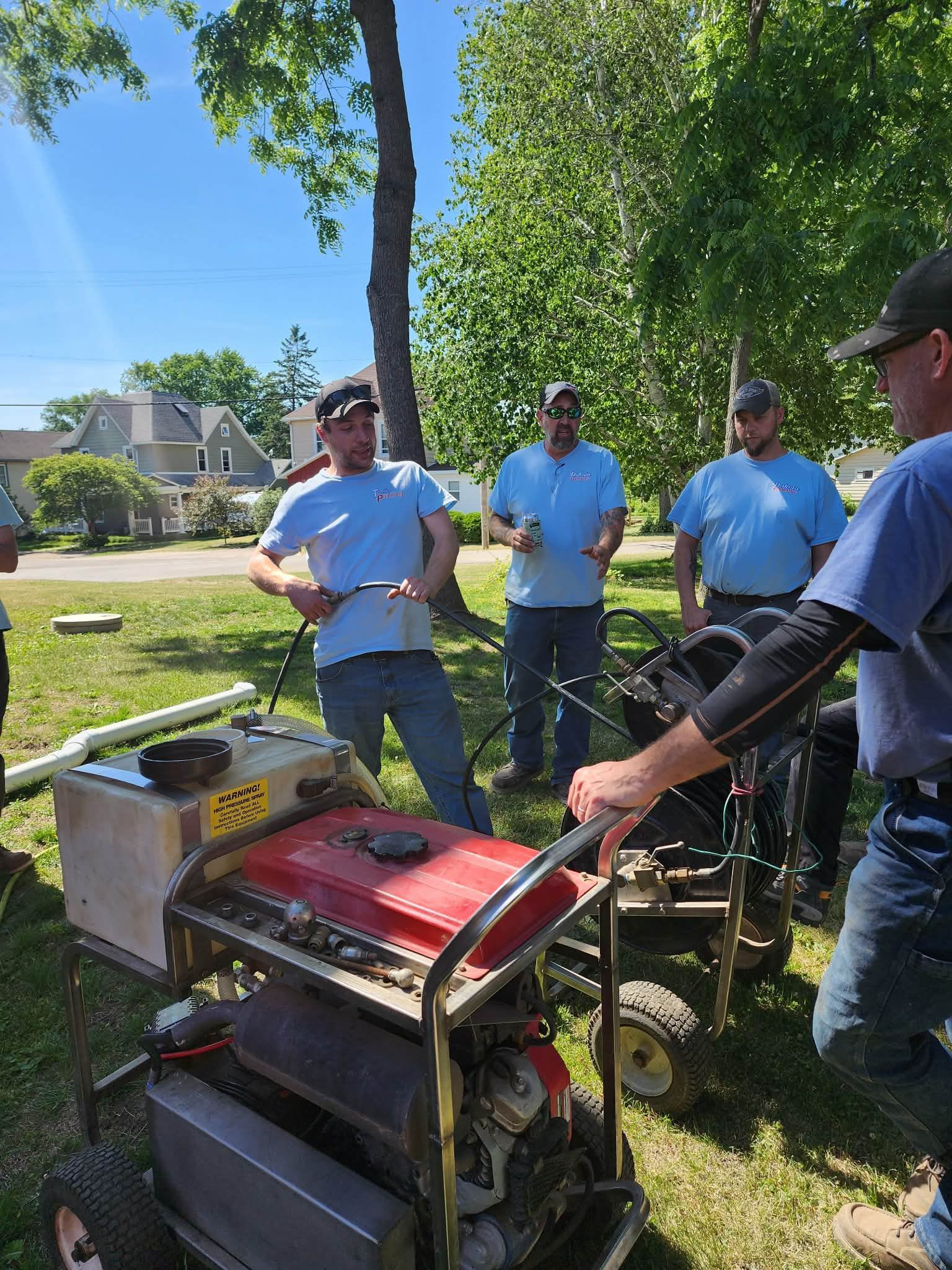People gathered around a red-and-black machine outdoors in a grassy yard on a sunny day
