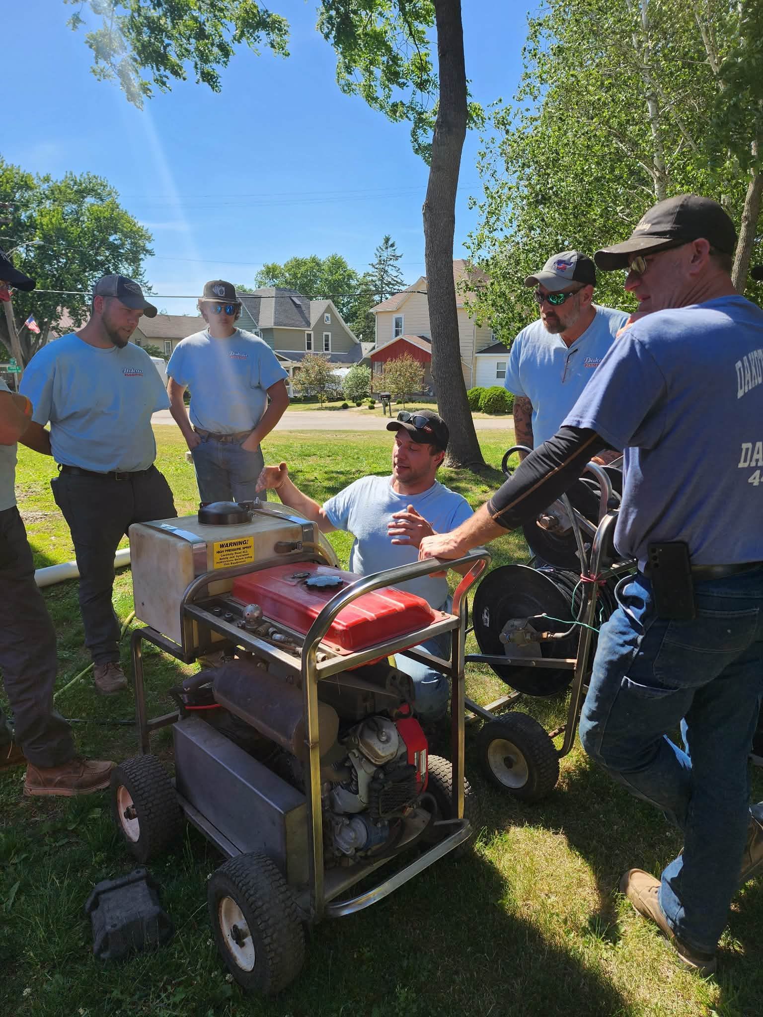 Group gathered around a small generator at a sunny outdoor event in a park-like yard.